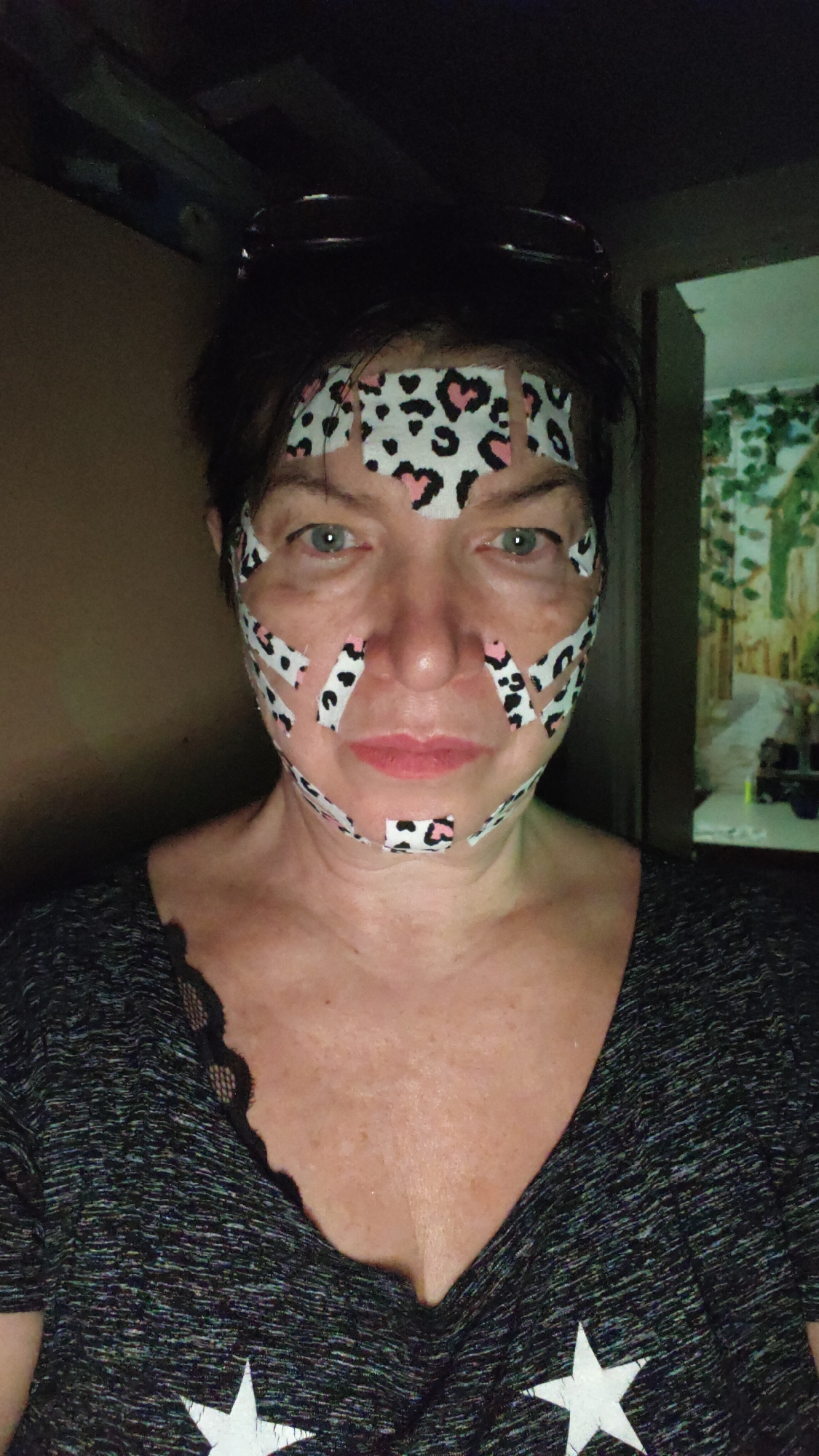 A medium-shot portrait depicts a woman looking directly at the camera in what appears to be an indoor, dimly lit setting. She has short, dark hair pulled back with sunglasses resting on her head. Her face is extensively covered with numerous small pieces of patterned tape, specifically white tape featuring black leopard spots and pink hearts. These tapes are strategically placed across her forehead, around her eyes, on her nose, cheeks, and chin, suggesting a facial aesthetic or beauty treatment. Her eyes are light-colored, appearing blue or green, and her expression is neutral.
She is wearing a dark, textured v-neck t-shirt, possibly grey or black, with a visible lace trim detail on the left shoulder. A large white star is partially visible on the lower right portion of her shirt. Her visible décolletage shows fair skin with some freckles or sunspots.
The background to her left is dark and indistinct. To her right, a brightly illuminated rectangular object displays a landscape scene with trees and a path, possibly a painting, a framed print, or a screen. The overall ambiance suggests a night-time or low-light environment within a home. The context given is Krasnoyarsk, Russia, though no specific visual cues within the image directly confirm this location. No discernible text is visible on the tape or clothing, only the star graphic.