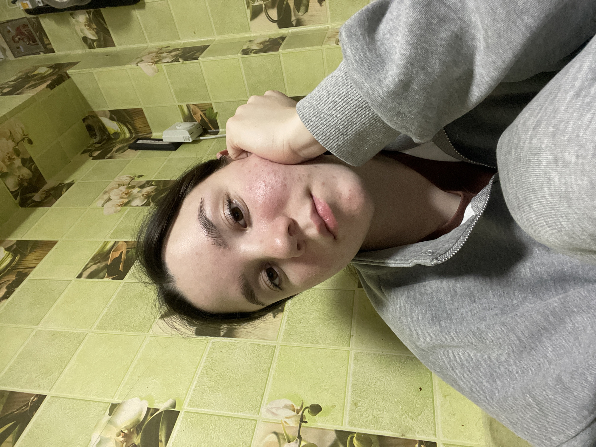 A young female subject, appearing to be in her late teens or early twenties, is depicted in a medium-close-up, looking directly at the camera. She has dark hair, dark eyes, and a neutral expression. Her skin exhibits visible redness and acne on her cheeks and chin. She is wearing a grey, ribbed, zip-up hoodie over a dark red undershirt. Her right hand is positioned with fingers resting against her right cheek and ear, supporting her head. The background consists of a wall covered in square tiles. Predominantly, these are textured light green tiles. Interspersed are decorative tiles featuring images of white orchids against a dark, possibly wood-patterned background. In the upper right section of the background, a small framed image, possibly a religious icon depicting two figures, is visible. Below it, a white electrical outlet or switch is mounted on the wall with a dark cable extending downwards. The scene takes place indoors, in the city of Ольховка, Russia.