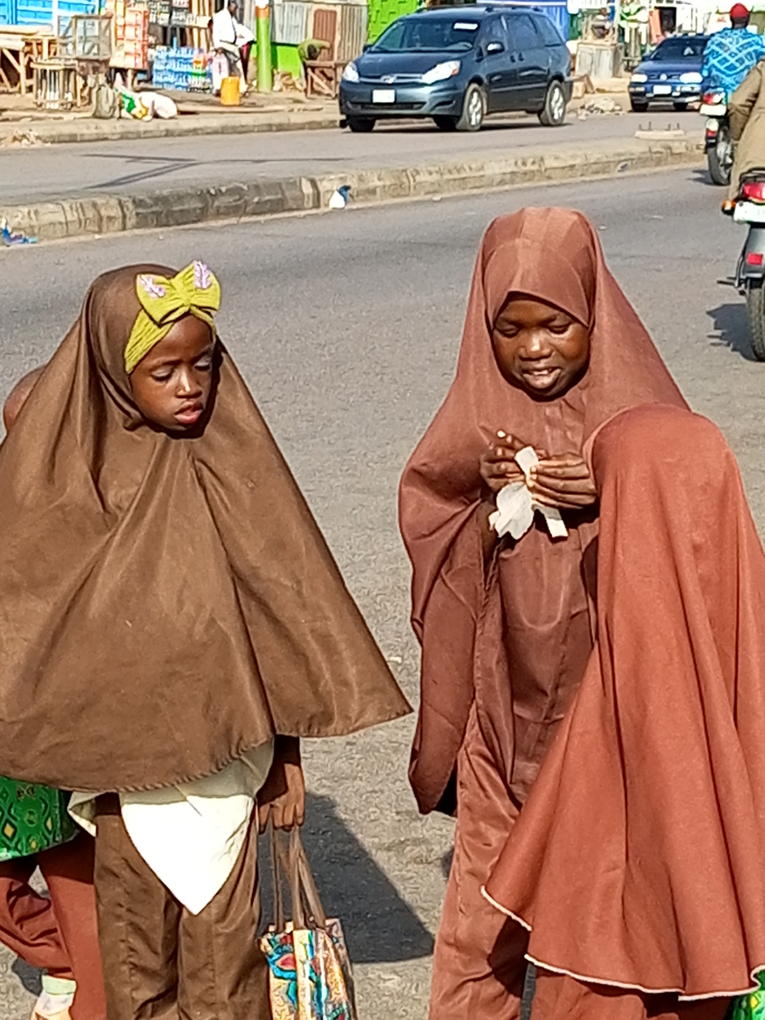 Three young girls in hijabs stand on a paved road. The girl on the left, wearing a brown hijab with a lime green bow, looks down. To her right, a girl in a brown hijab is holding a white tissue. Another girl, partially visible on the right, is also wearing a brown hijab. The scene appears to be a street in Tempure, Nigeria, with a road, sidewalk, and shops visible in the background. Cars and a motorcycle are on the road, suggesting a typical daytime scene. The lighting indicates it is daytime.