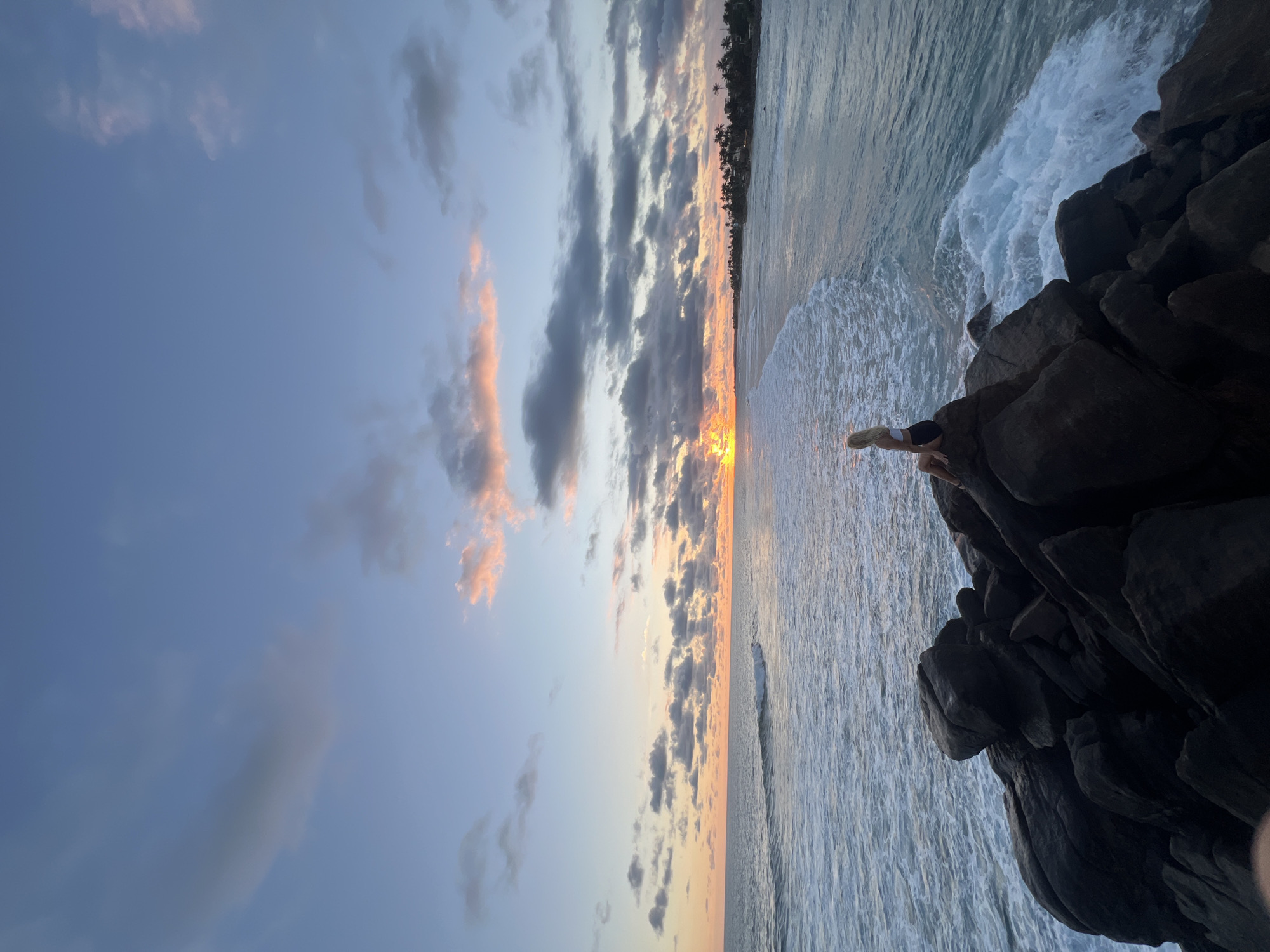 The image shows a sunset scene in Goviyapana, Sri Lanka. The sky is a mix of blue and orange with scattered clouds. Below, the ocean stretches to the horizon where the sun sets, creating a golden glow. A person is sitting on rocks at the edge of the shore. Waves crash around the rocks. Palm trees are visible along the coastline in the distance.