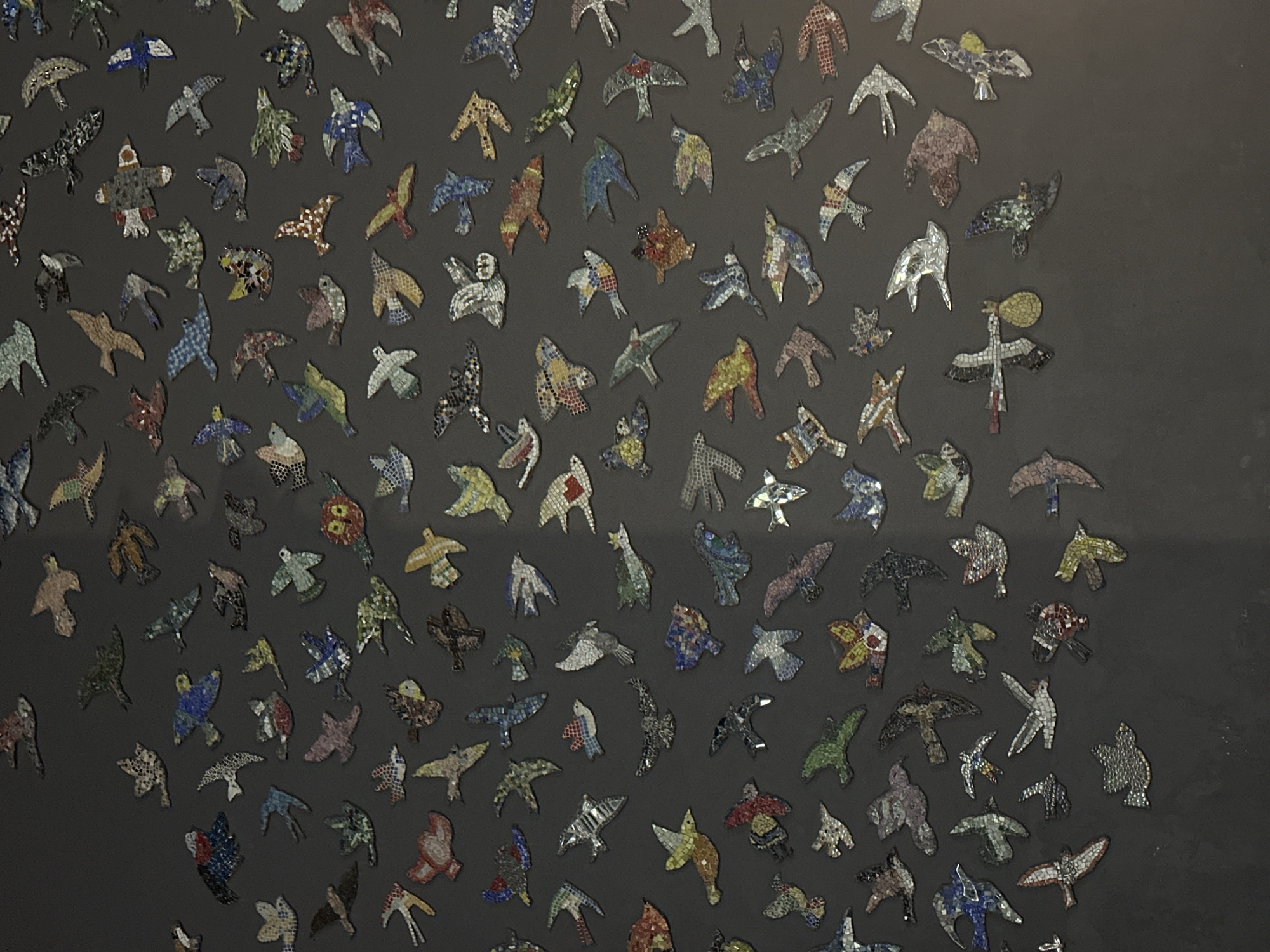 The image displays an artistic installation featuring a multitude of small, bird-shaped mosaics adhered to a dark gray wall, located in Novosibirsk, Russia. Each mosaic bird is uniquely crafted from numerous small, irregularly shaped fragments of various materials, likely glass, ceramic, or mirror, presenting a rich tapestry of colors and textures.
The birds exhibit a wide spectrum of hues, including blues, greens, reds, yellows, browns, and whites, with some incorporating reflective or iridescent pieces. They are depicted in diverse poses and orientations across the entire surface, suggesting a dynamic flock in flight or a scattered gathering. Some birds have outstretched wings as if soaring, while others appear more compact or perched. The mosaic technique is clearly evident, highlighting the intricate assembly of tiny tesserae that form each avian silhouette.
The uniform dark gray background provides a stark contrast, allowing the vibrant and detailed mosaic birds to stand out prominently. The lighting appears even, indicating an indoor setting or a controlled display environment. The artwork evokes a sense of movement, diversity, and artistic craftsmanship. No text is visible within the image.
