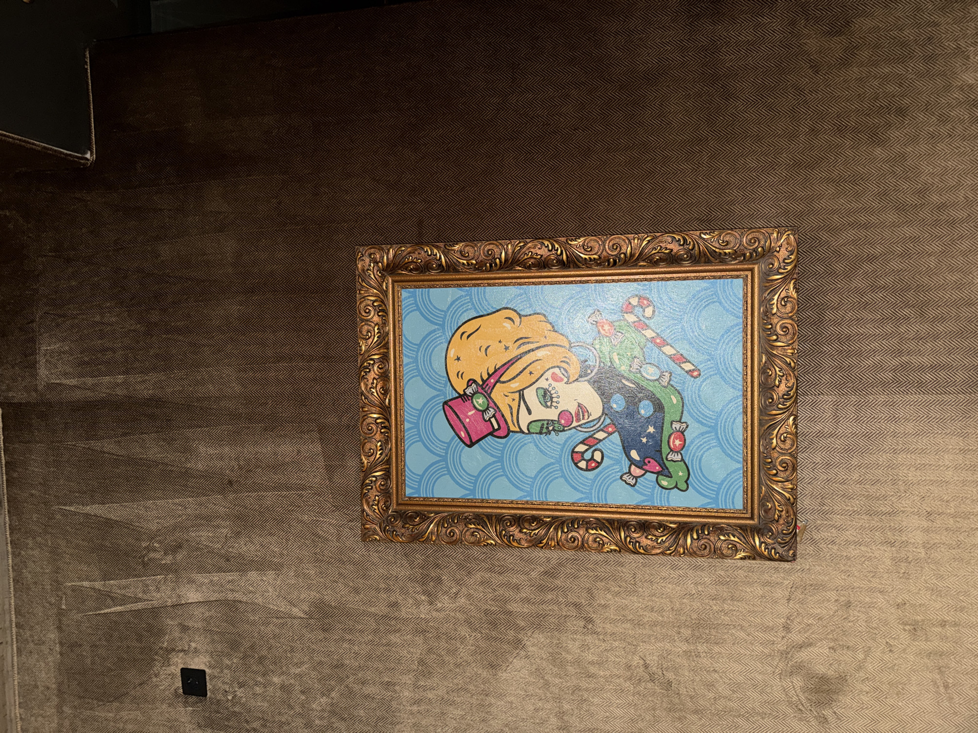 A framed, whimsical painting hangs on a textured, herringbone-patterned wall. The painting features a cartoon-style figure with blond hair, a pink hat, and candy cane details against a light blue, patterned background. The frame is ornate and gold-colored. The wall is dark brown with lighter areas and some dark spots near the bottom left, possibly a power outlet. The location in Sofia, Bulgaria is not evident from the image.
