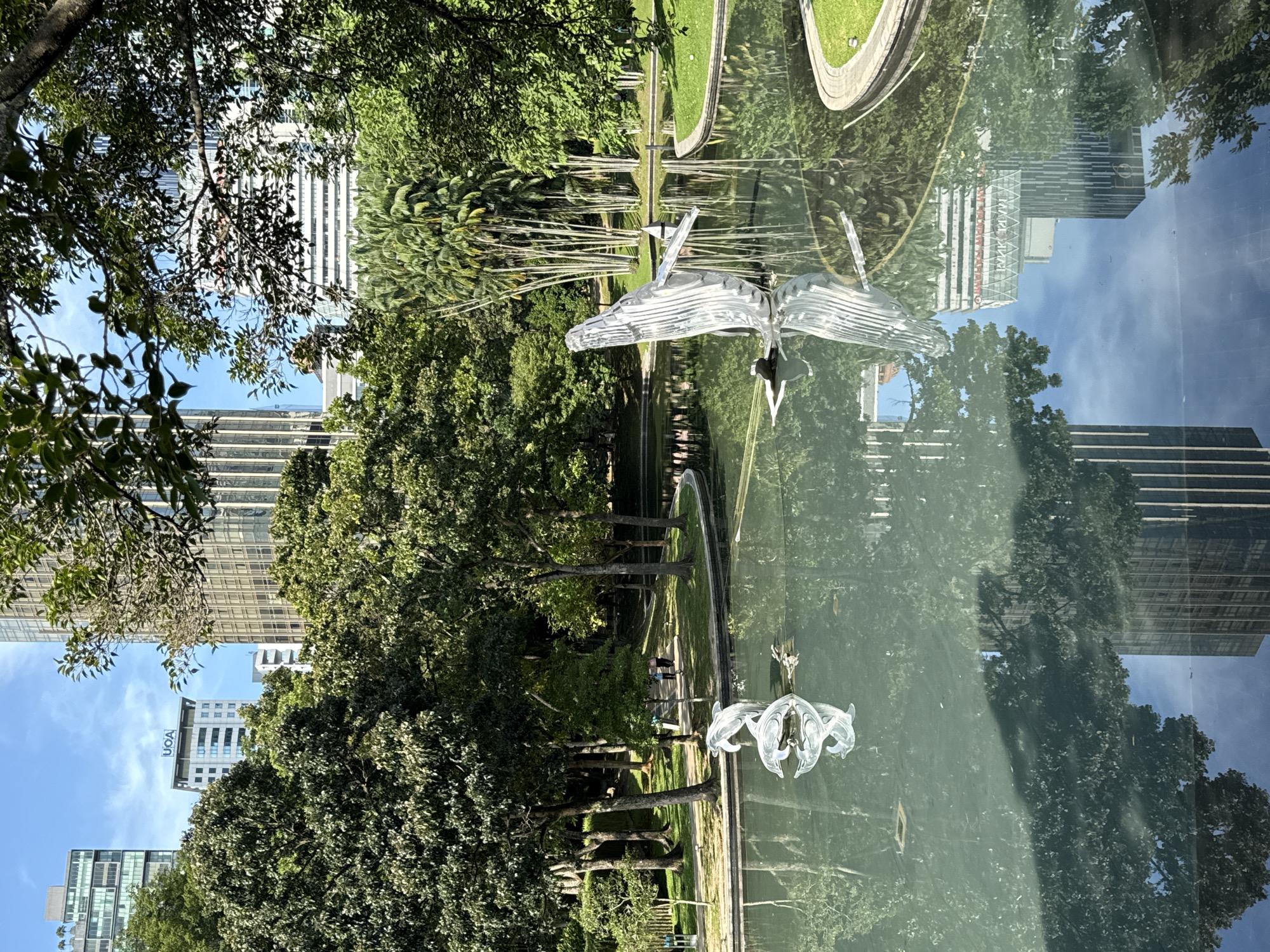 The image depicts a park scene in Kuala Lumpur, Malaysia. The view is reflected in a still body of water, possibly a pond or pool. Trees line the edges of the water, with a walking path visible. A translucent sculpture of a bird, possibly made of glass or plastic, is positioned in or above the water, reflecting in the surface. In the background, modern high-rise buildings are visible, some reflecting in the water as well. The sky is clear and blue. A couple of people can be seen on the path. The overall impression is one of a tranquil urban park setting.
