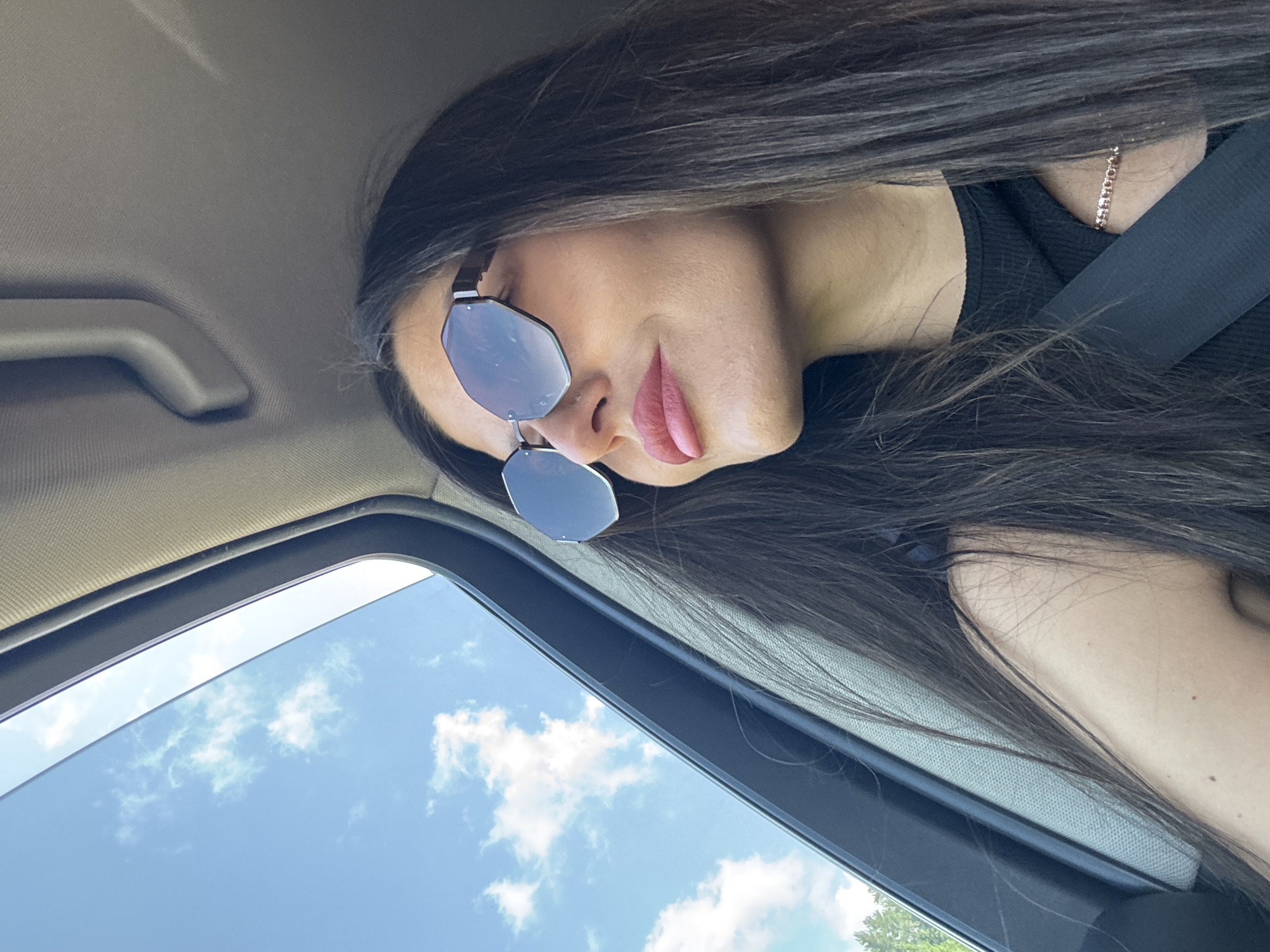 The image shows a woman with long, dark hair and sunglasses, looking out of a car window. She is wearing a black top and a seatbelt. The sky is blue with some clouds visible. The scene appears to be a daytime shot, possibly during a car journey. There is no discernible location information within the image that can confirm it is in Olkhovka, Russia.