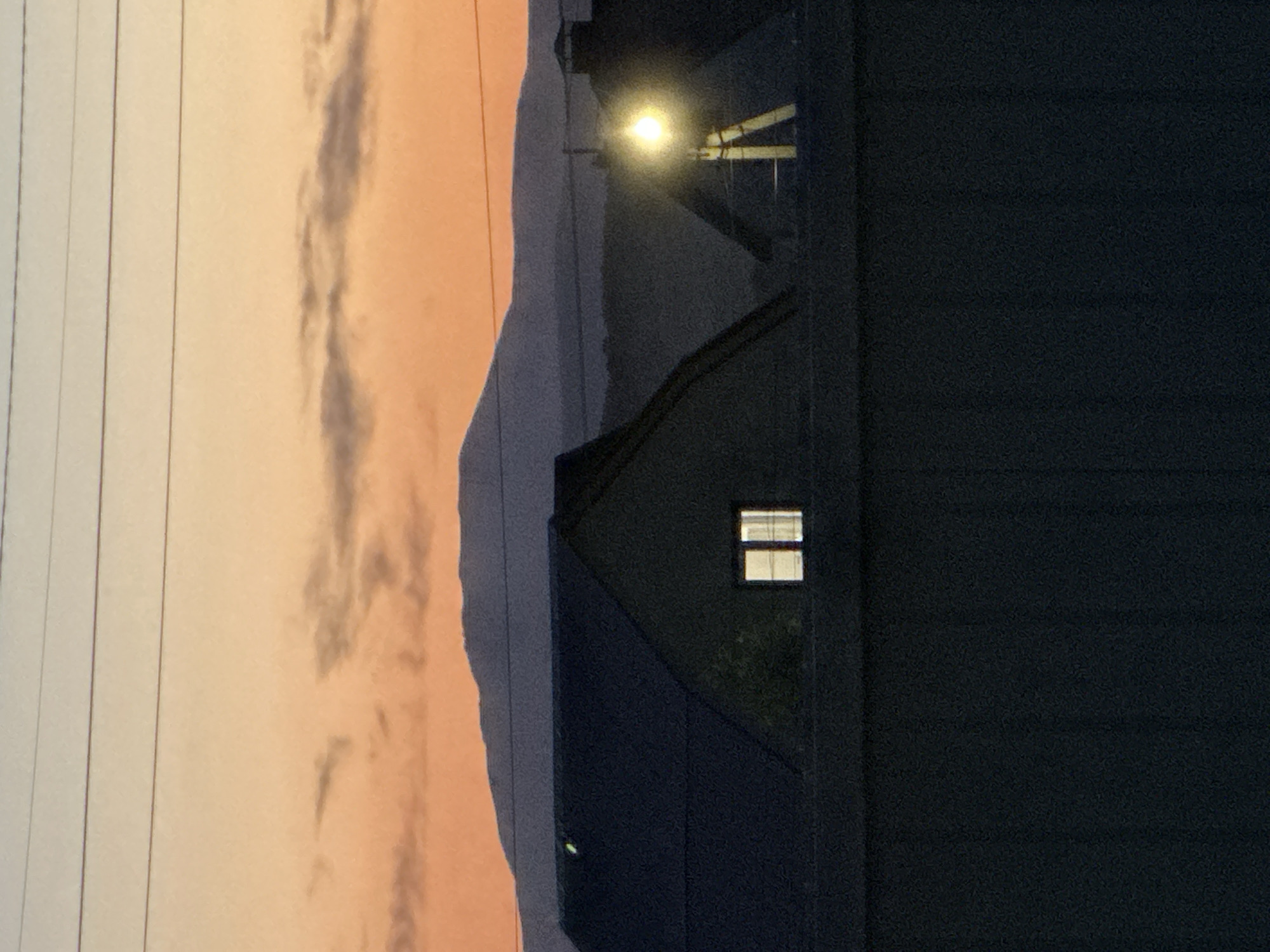 The image depicts a building with a lit window at dusk, possibly located in the city of Unknown, Unknown. The sky is a blend of orange and grey, suggesting either sunset or sunrise. Utility lines are visible on the left side of the frame. A streetlight is faintly visible in the background, suggesting the presence of other buildings.