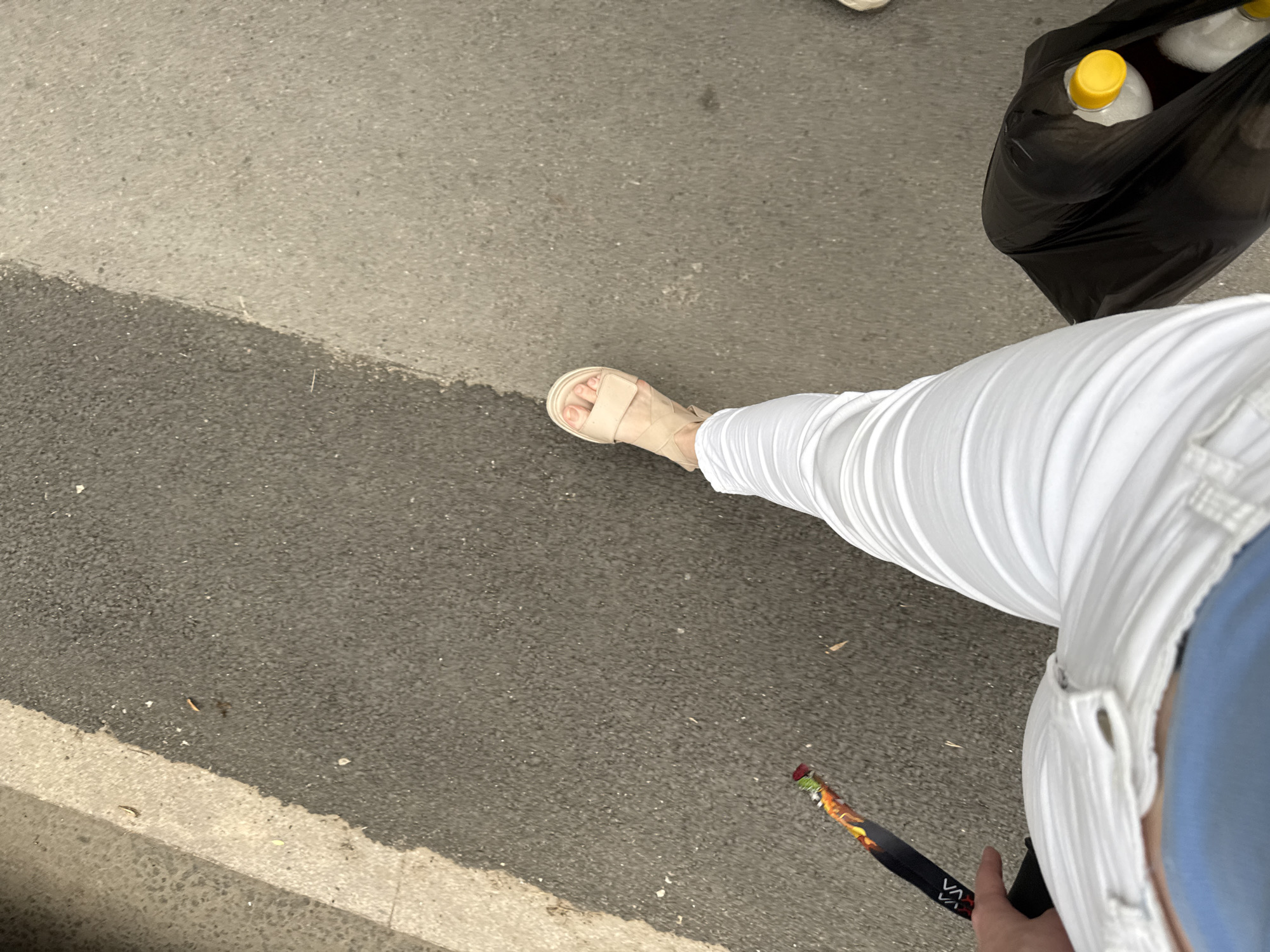 The image shows a person's legs and feet as they walk on a paved surface, possibly a sidewalk or street. The person is wearing white, wide-legged pants and light-colored sandals. In their left hand, they are holding a black object with a colorful, patterned design. To the right of their legs, a black plastic bag is visible, containing at least two white bottles with yellow caps. The ground appears to be asphalt with some lighter-colored concrete or stone sections. The environment suggests an outdoor urban setting. No specific location in Almaty, Kazakhstan, can be identified from the visual information alone.