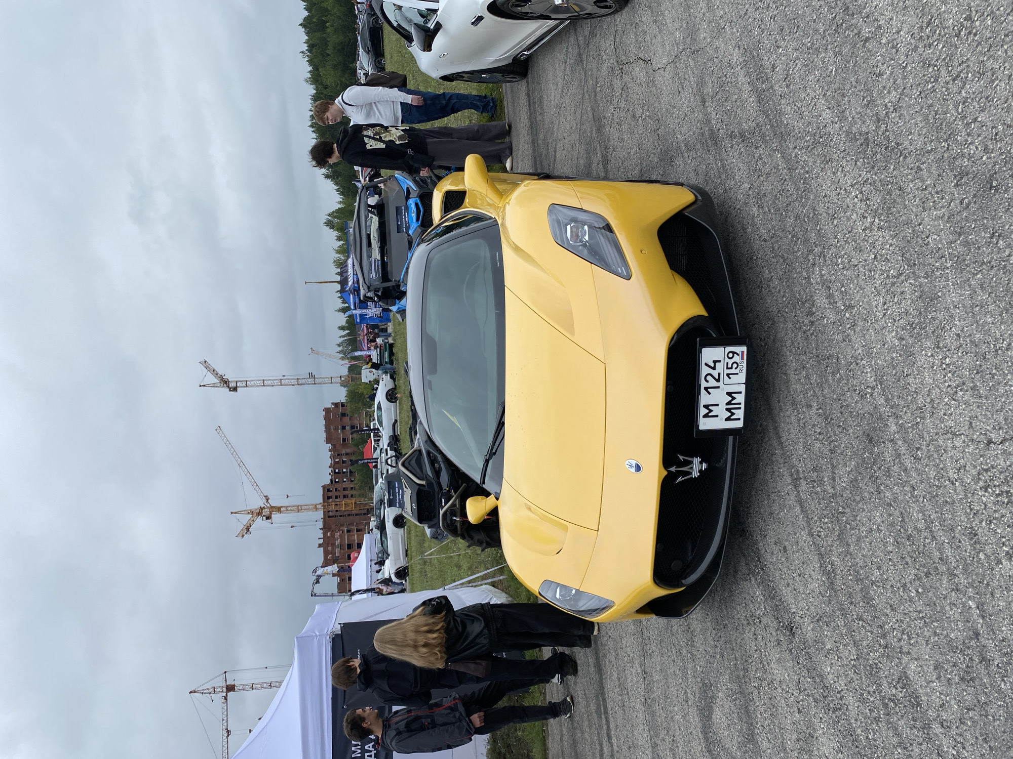The image captures an outdoor event, likely a car show or exhibition, under a cloudy sky. In the foreground, a bright yellow Maserati sports car is prominently displayed. The car's license plate, visible and partially obscured, reads "M 124 MM 159" with "RUS" below. The car is positioned on an asphalt surface. Behind the Maserati, several other vehicles are visible, including a white sports car on the right and a blue off-road vehicle. A group of people are gathered near tents on the left side of the frame, some observing the vehicles. In the background, construction cranes and partially built structures suggest ongoing development in the vicinity. The presence of the "RUS" on the license plate indicates the location is in Russia. While the specific city of Perm is not explicitly stated in the image, the context of an outdoor event with a variety of vehicles and construction in the background are the key elements of the scene.