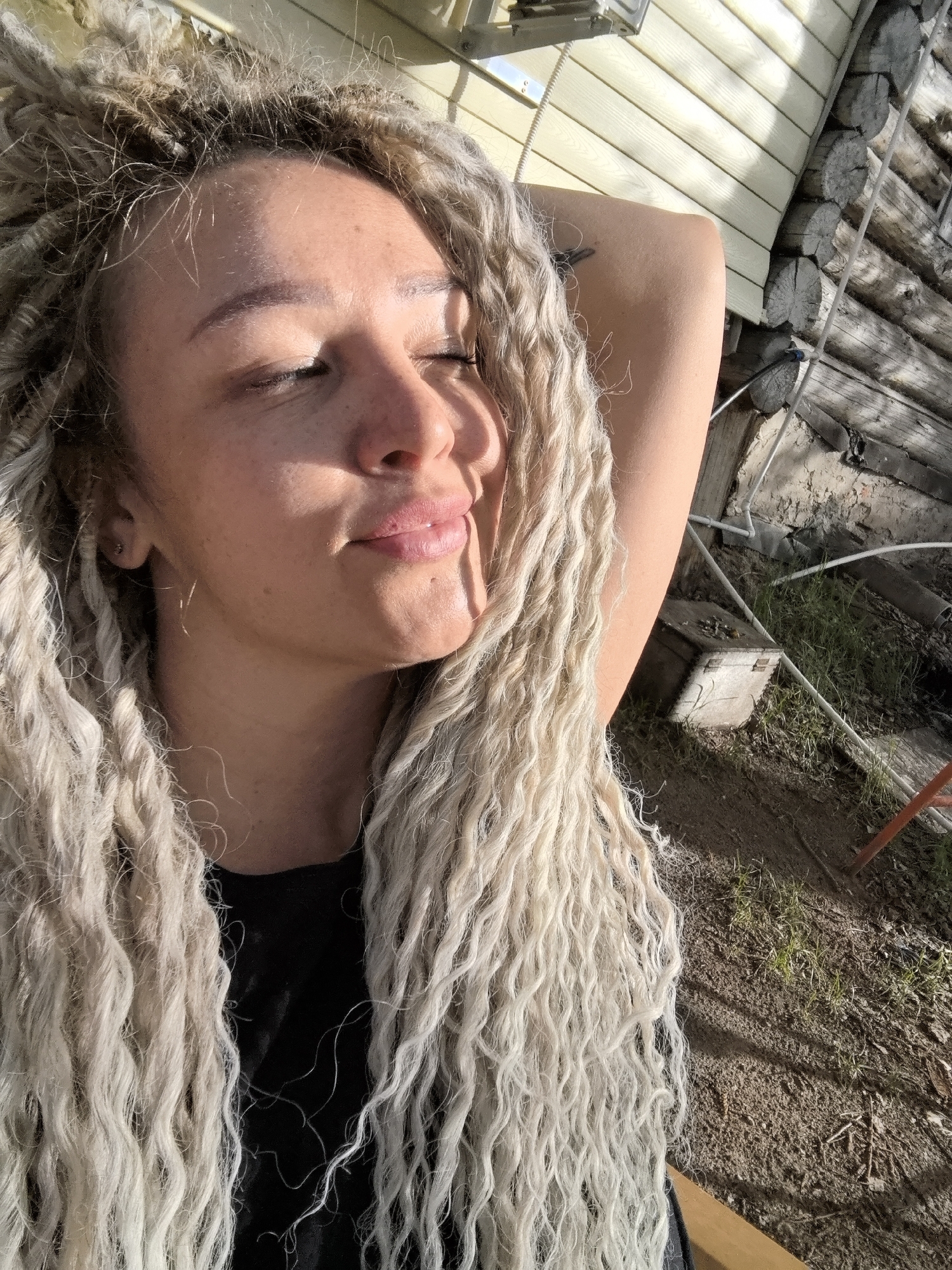 A fair-skinned woman with long, light-colored dreadlocks is facing the sun with her eyes closed. She is wearing a black top. Her right arm is raised behind her head. The background includes a wooden structure with light-colored siding and a log wall, a dirt ground with sparse grass, and some pipes. The location is in a partially sunny outdoor area, presumably in Noviy, Russia.