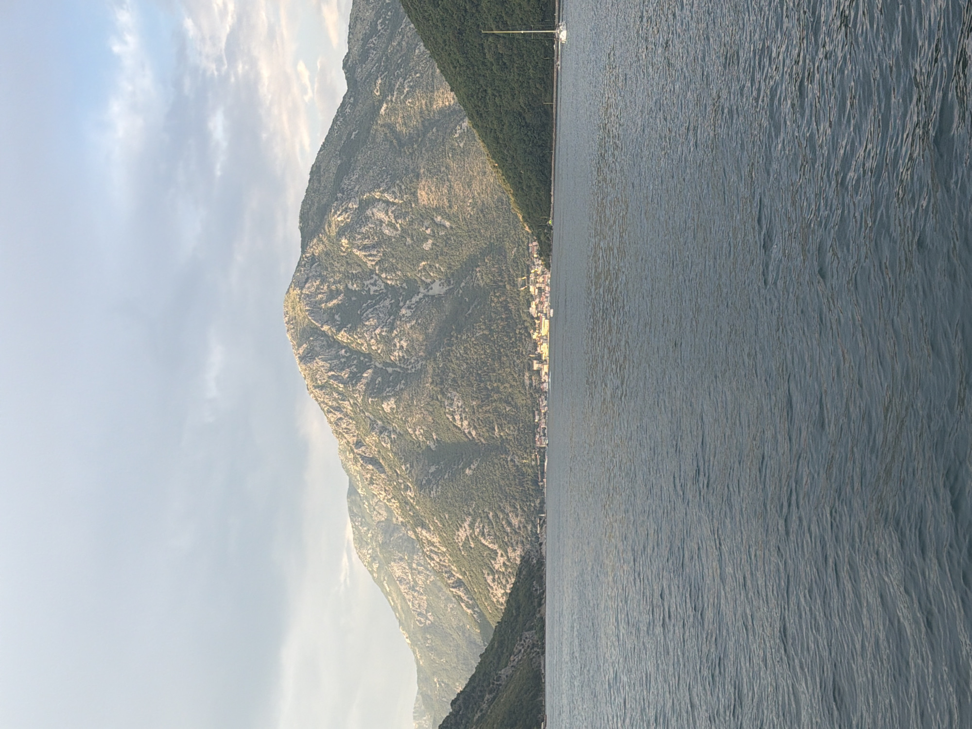 A wide shot captures a serene natural landscape dominated by water and mountains. In the foreground, a body of calm, dark blue water stretches across the frame, with subtle ripples indicating a gentle movement. Behind the water, a picturesque town is nestled at the base of towering, rugged mountains. The mountains, partially covered in dense green foliage on their lower slopes, rise dramatically, their rocky peaks catching the soft light of what appears to be late afternoon or early morning. The sky above is a soft grey, with scattered clouds creating a diffused light that casts a gentle glow on the scene. To the right of the frame, a solitary white sailboat with its mast extended is a subtle focal point against the vastness of the water and mountains. The overall impression is one of tranquility and natural beauty, with the town providing a human element to the majestic scenery.