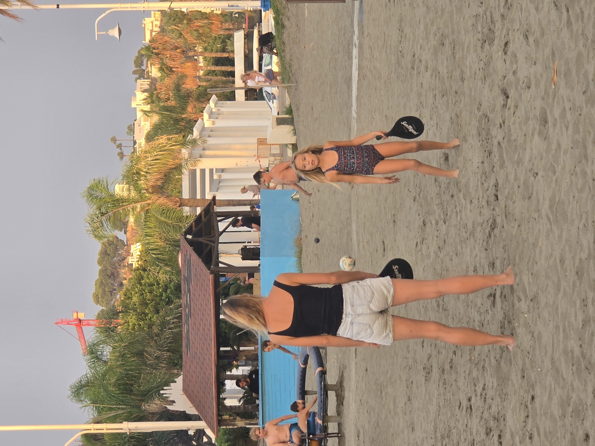 This image captures a beach scene in Málaga, Spain, likely during the late afternoon or early evening due to the warm, diffused light. In the foreground, two individuals are engaged in a beach paddle game. A woman with blonde hair, wearing a black tank top and denim shorts, stands facing away from the camera, holding a paddle. In front of her, a young girl in a patterned swimsuit is also holding a paddle, facing the woman. A beach ball is visible in the sand between them.
The background reveals a lively beach environment. A blue-colored structure, possibly a bar or changing rooms, is present, with several people near it. Some individuals are seated on a bench-like structure, while others stand around. Palm trees and other tropical vegetation are scattered throughout the scene, characteristic of a coastal area. In the distance, buildings and structures are visible on a hillside, suggesting an urban backdrop. A red construction crane is prominently visible above the trees in the far background. The ground is sandy, typical of a beach, with footprints and slight undulations in the sand. The sky is overcast with a soft, diffused light.