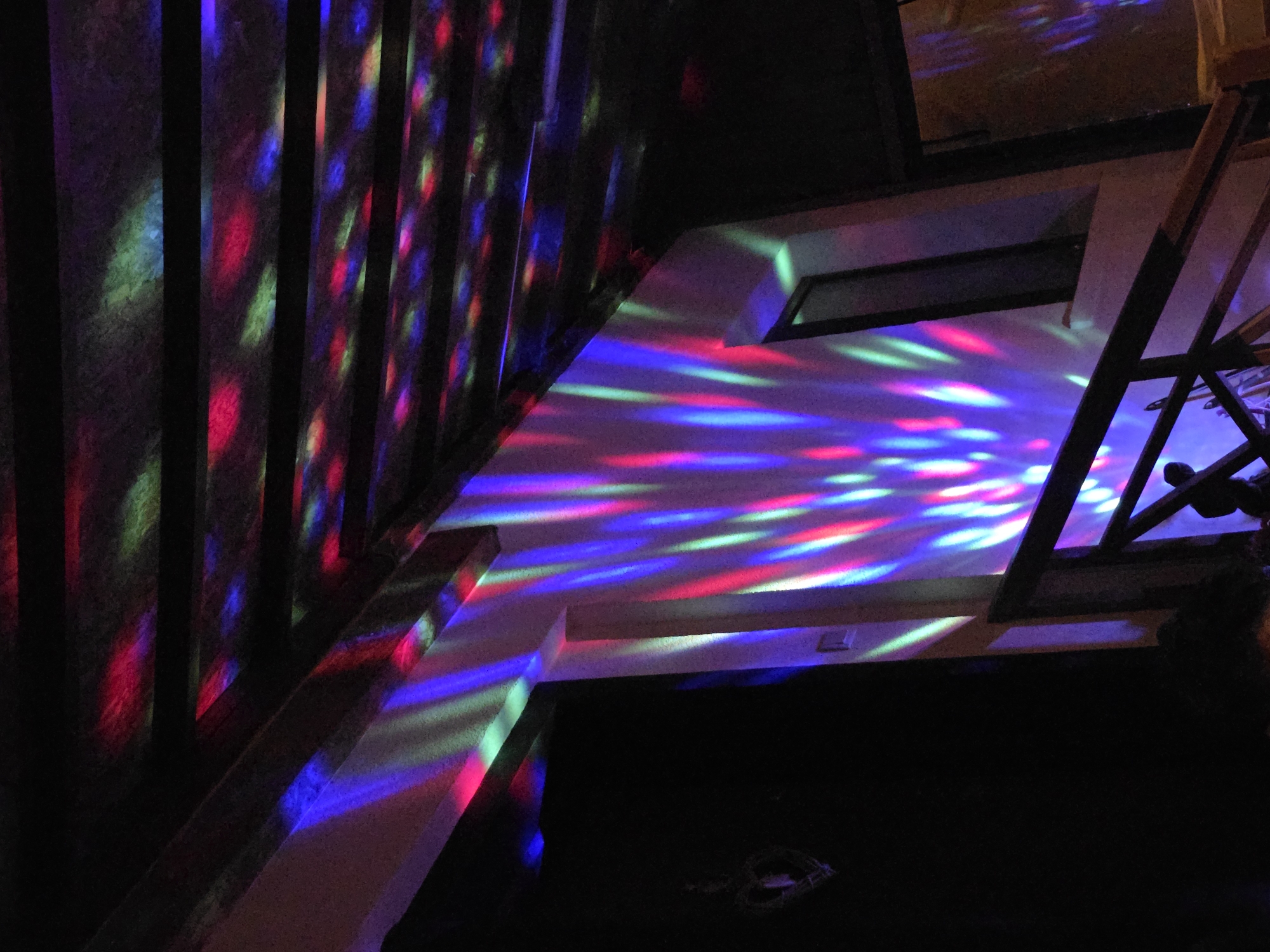 A dark, interior shot showing a staircase with colorful, patterned light projections. The light patterns are composed of red, blue, and green streaks, appearing on the stairs and walls. The scene includes structural elements of a staircase, such as railings and steps, as well as portions of walls. There are no people visible. The location, Pokrayna, Bulgaria, cannot be independently confirmed from the image content.