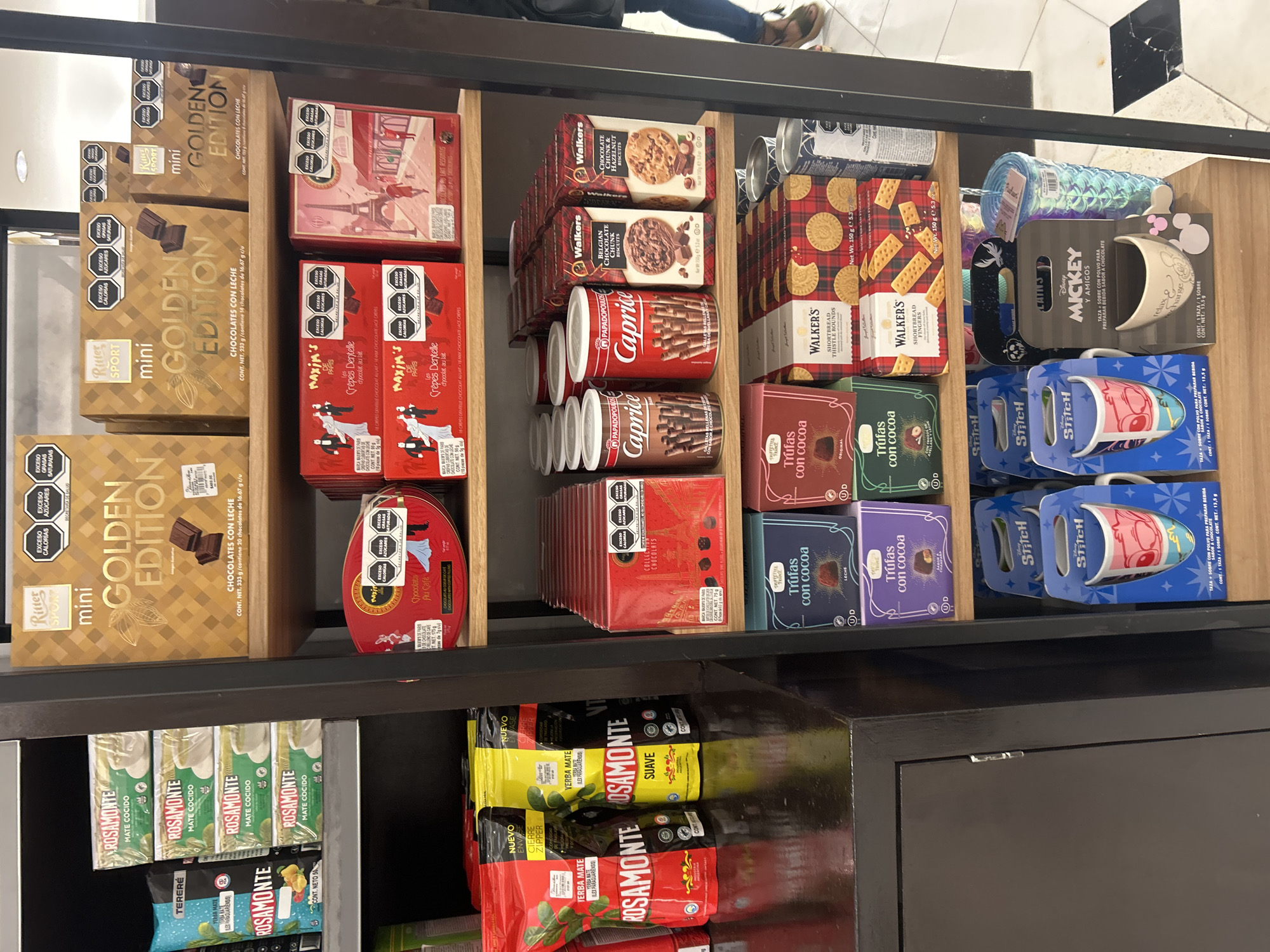 A shelf displays an assortment of packaged food products. The location is a store in San Bernardino Tlaxcalancingo, Mexico.
Top Shelf:
- "Mini Golden Edition" Ritter Sport chocolates in gold packaging.
- Boxes of "Maxim's Paris" Crepes Dentelle chocolate cookies, in red packaging.
- Walkers biscuits.
- Red and white Caprice wafer rolls in tins.
- Boxes of "Trufas con Cocoa" chocolates in various colors.
- Disney-themed mugs of Stitch and Mickey.
Bottom Shelf:
- "Rosamonte Mate Cocido" tea bags.
- "Rosamonte" yerba mate tea in various packages, including "Suave".
The shelf appears to be part of a display inside the store, and the overall impression is of a retail setting where these items are available for purchase.