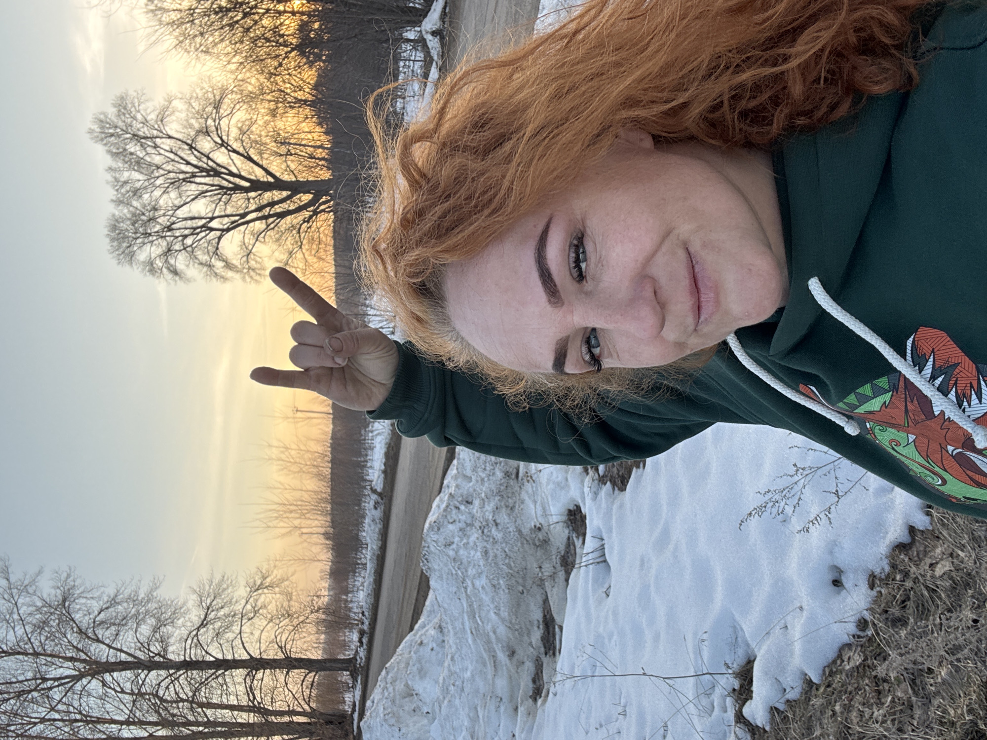 A selfie taken outdoors in what appears to be a rural or semi-rural area with snow on the ground. A woman with red hair is the primary subject, wearing a dark green hooded sweatshirt. She is making a "rock on" hand gesture. In the background, there's a road, patches of snow, bare trees, and a golden or sunset-lit sky. No information about the specific location (Noviy, Russia) can be confirmed from the image.