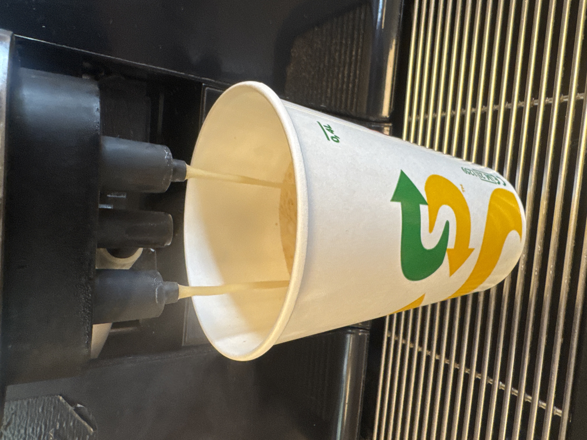 In an indoor setting, likely a Subway restaurant, a white paper cup with the Subway logo and branding is positioned beneath a black coffee machine. Two streams of light brown liquid are pouring from the machine into the cup, which is approximately half-filled. The cup rests on a black metal grate. The background is dark, with the coffee machine dominating the upper portion of the image. The scene is static, with no people or animals present.