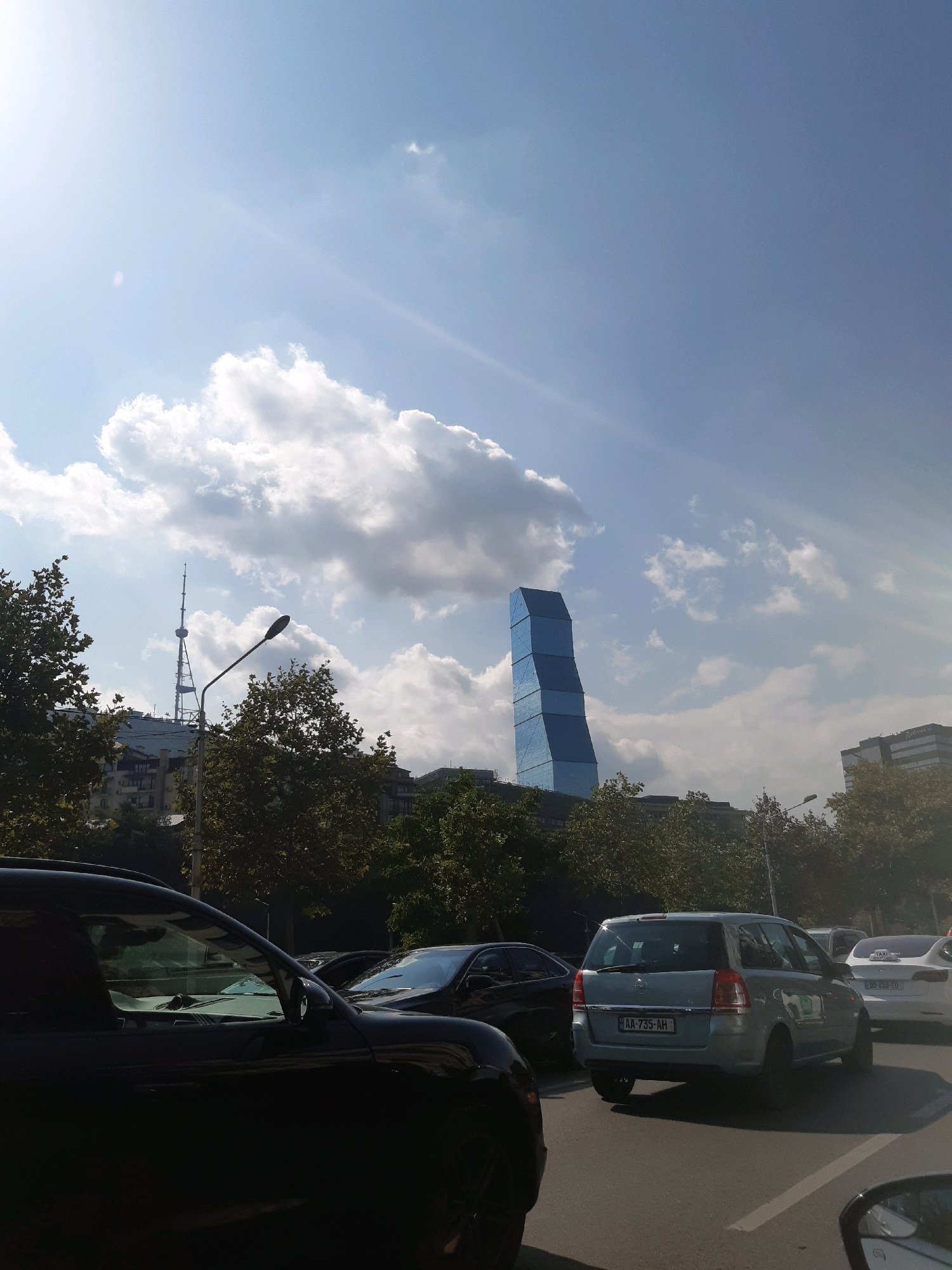 The image captures a street scene in Tbilisi, Georgia, with a distinctive blue, twisted skyscraper dominating the skyline. The sky above is a bright blue with scattered white clouds, and the sun casts a slight glare. In the foreground, several cars are visible, including a dark-colored SUV on the left, a light blue station wagon in the center with license plate AA 735 AH, and a white sedan on the right. Trees line the street, partially obscuring buildings in the mid-ground, one of which appears to be a television tower. The overall impression is a sunny day in a modern urban environment.