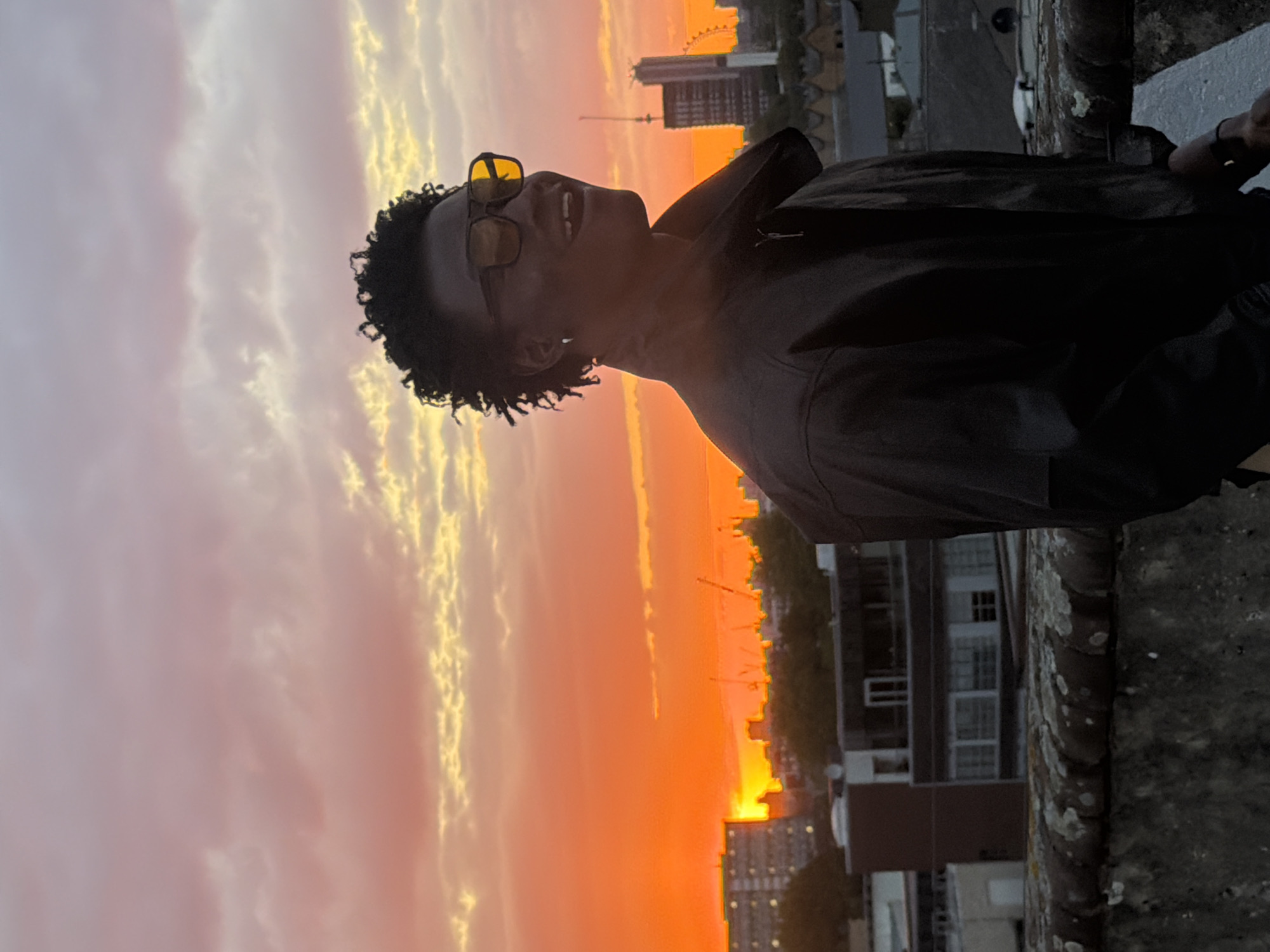 A medium shot shows a dark-skinned person with dark, curly hair, wearing yellow-tinted glasses, and a black jacket, turned to the left against a vibrant sunset. The person is standing on a rooftop or ledge made of gray stone. In the background, the London skyline is visible, including buildings and the London Eye. The sky transitions from a light, cloudy pink to a bright orange near the horizon. The person is smiling slightly, with their head tilted back. The overall lighting is low, accentuating the sunset.