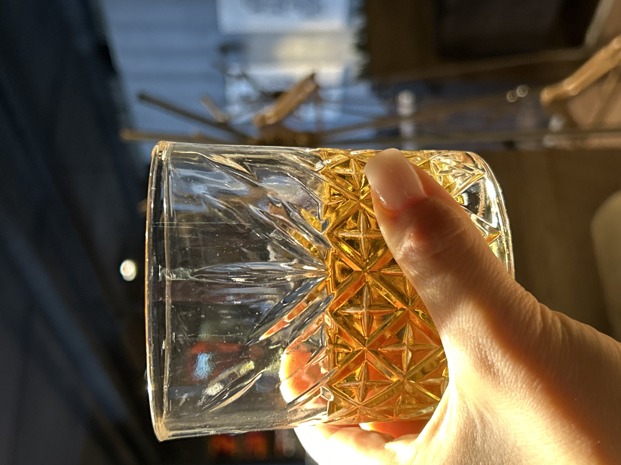This close-up image, captured in Chaikovsky, Russia, features a person's hand holding a clear, patterned glass. The hand, with a neatly manicured fingernail, grips the glass from below, its fingers partially covering the lower section. The glass has two distinct textures: one half, held by the hand, features an intricate, faceted geometric pattern which glows with a rich golden-amber hue, likely due to the interaction of light with the glass's texture and the golden liquid it contains. The other half of the glass is smooth and clear, with subtle vertical etched lines. The liquid inside the glass appears to be a golden-amber color. The background is softly blurred, showing dark, indistinct shapes on the left (possibly fabric or a curtain) and warmer, abstract light reflections and linear elements on the right, suggesting an indoor setting. The overall lighting is warm and somewhat dramatic, hinting at an evening or late afternoon ambiance. There is no visible text in the image.