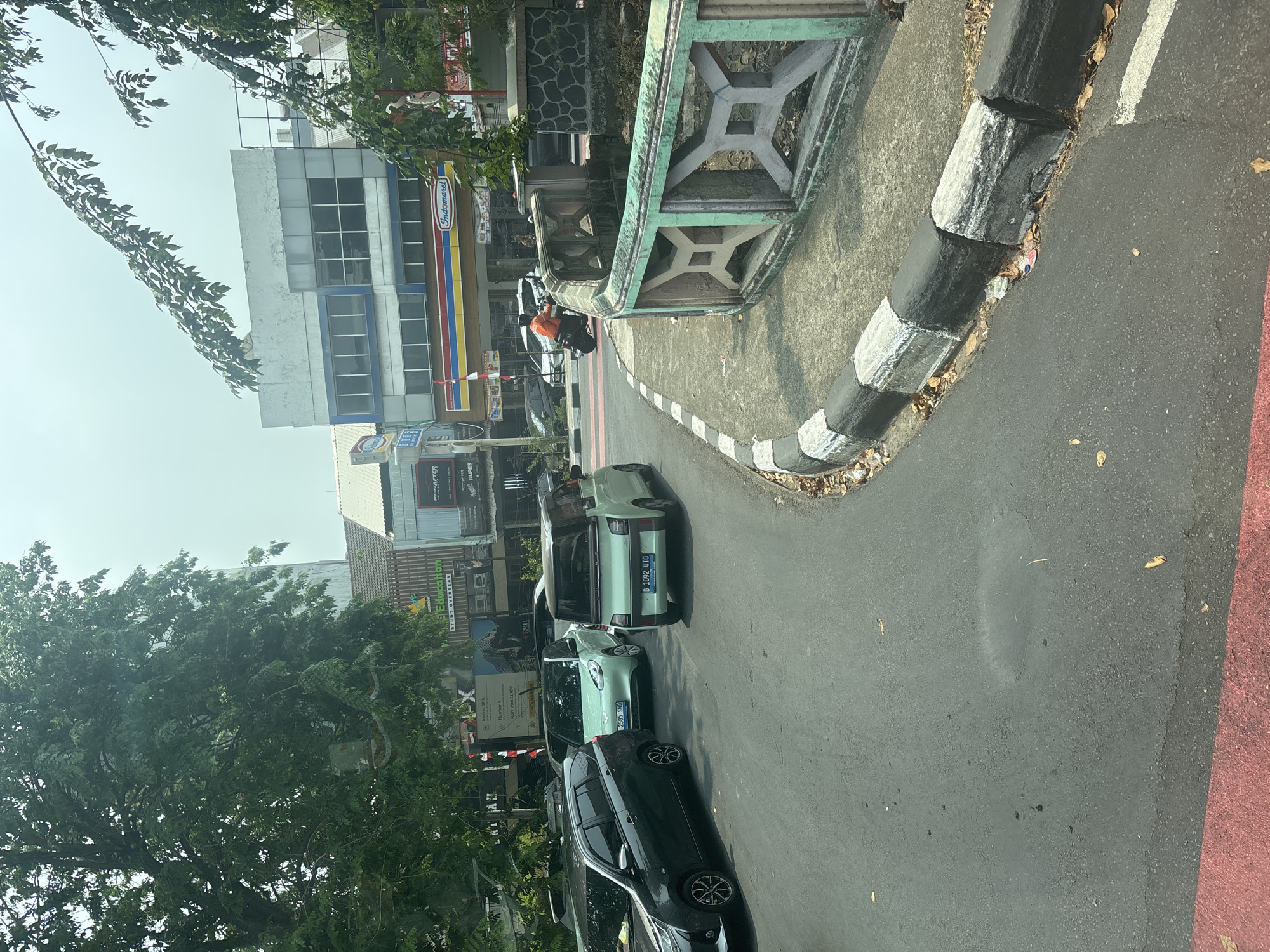 The image shows a street scene in North Jakarta, Indonesia, under a hazy sky. A dark grey minivan is parked on the left side of a paved road, facing away from the viewer. To its right, a light green car with a black roof is also parked, facing the same direction. Further ahead, a similar light green vehicle, a small electric car, is driving on the road. Behind it, a motorcycle with a rider wearing an orange jacket is also moving.
On the right side of the road, there is a sidewalk with a decorative, light green and grey patterned railing. Beyond the railing, there appears to be a stone wall. In the background, several multi-story buildings are visible. One building has a prominent sign that reads "Indomaret" with its characteristic red, yellow, and blue stripes. Another building displays a sign with "Education" and "Study Overseas" suggesting it's an educational institution. The buildings are partially obscured by lush green trees with dense foliage. The road has white lane markings, and a pedestrian crossing with red and white stripes is visible in the distance. Debris, including leaves and some litter, is scattered along the edge of the sidewalk and road.