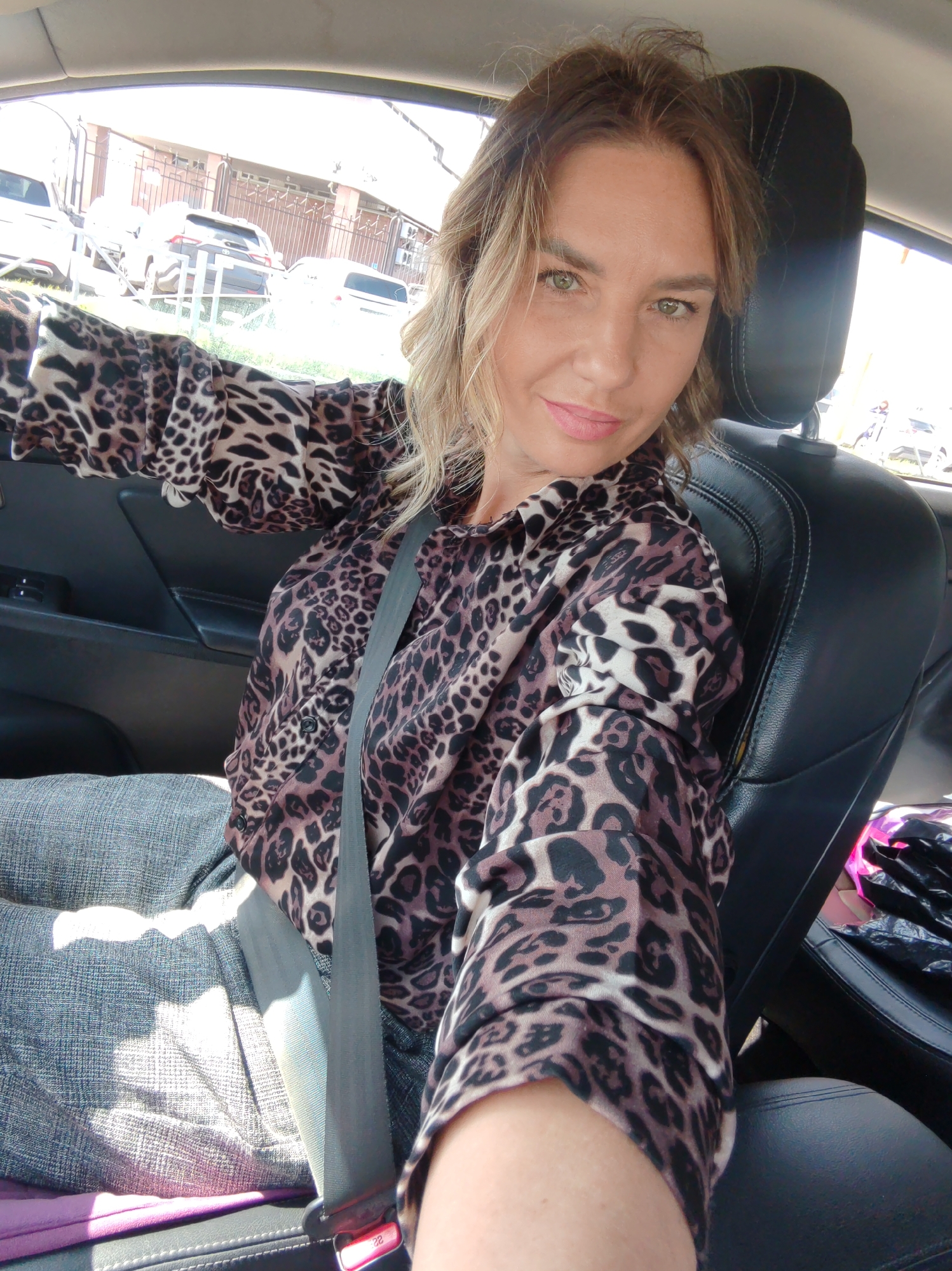 A woman is captured in a selfie taken inside a car during what appears to be a bright, sunny day. She is the central subject, positioned in the driver's seat, looking directly at the camera with a neutral to slightly pleasant expression. Her hair is medium-length, wavy, and has light brown or blonde highlights. She has green eyes and is wearing light pink lipstick.
She is dressed in a distinctive leopard print long-sleeved shirt and gray trousers, and a gray seatbelt is fastened across her chest and lap. Sunlight streams into the car, creating bright highlights on her clothing and the car's interior. The seatbelt buckle displays the number "22" in red.
Through the car's window, an outdoor scene is visible. Several vehicles, including SUVs, are parked outdoors. Behind the parked cars, there is a building made of dark reddish-brown bricks, fronted by a black metal fence. Some green foliage is also discernible in the background. The car's interior features black leather seats and a dark gray door panel. The overall scene suggests an ordinary moment of travel or waiting in Petropavlovsk-Kamchatsky, Russia.