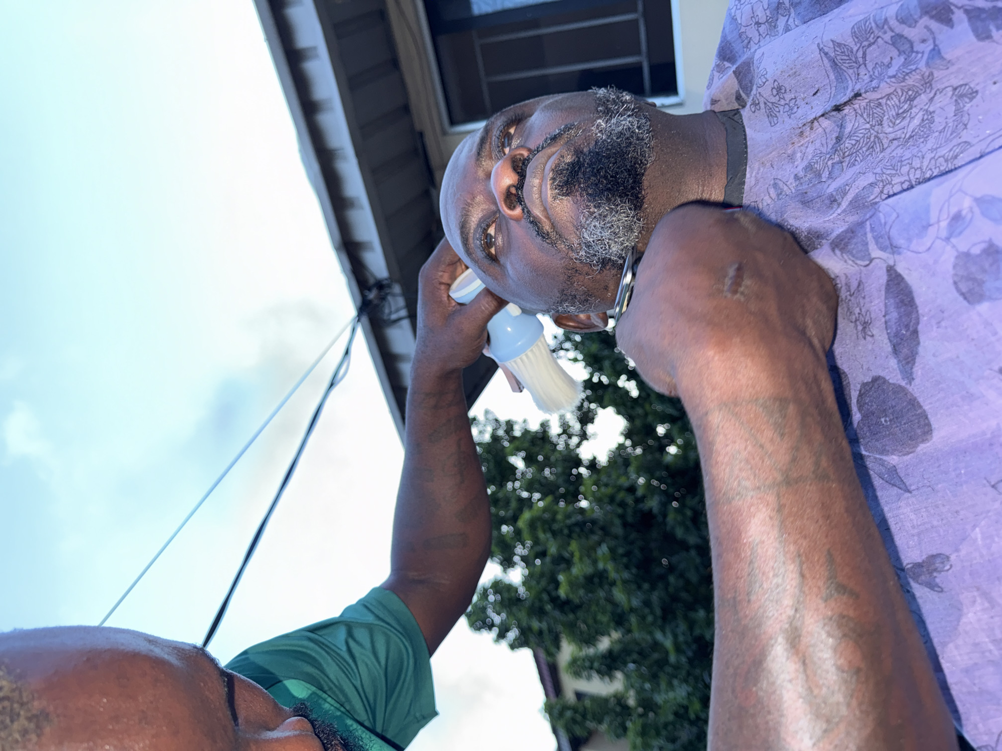 A low-angle shot captures a man with dark skin and a salt-and-pepper beard having his beard dusted with a small brush by another person. The man receiving the dusting is wearing a patterned purple shirt and silver earrings. The individual dusting is wearing a green shirt and has tattoos on their arm. The background includes a section of a building, some visible wires, trees, and a cloudy sky. The location is believed to be Ado, Nigeria, but no specific landmarks confirm this.