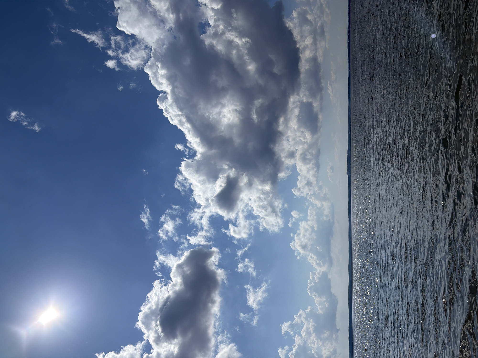 The image depicts a bright, sunny day with a vast expanse of blue sky filled with fluffy white and grey clouds. The sun, a brilliant white orb, is prominently visible in the upper left quadrant, casting a glare and lens flare across the sky. To the right of the sky, a body of water stretches to the horizon, its surface rippled and shimmering with sunlight. The water appears dark, possibly reflecting the sky or being deep. A faint, dark line on the horizon suggests land or trees in the distance. The overall scene evokes a sense of a clear day, likely midday or afternoon, with a calm and expansive natural setting. There is no visible text in the image. The orientation of the image is landscape with the sky occupying the left half and the water occupying the right half.