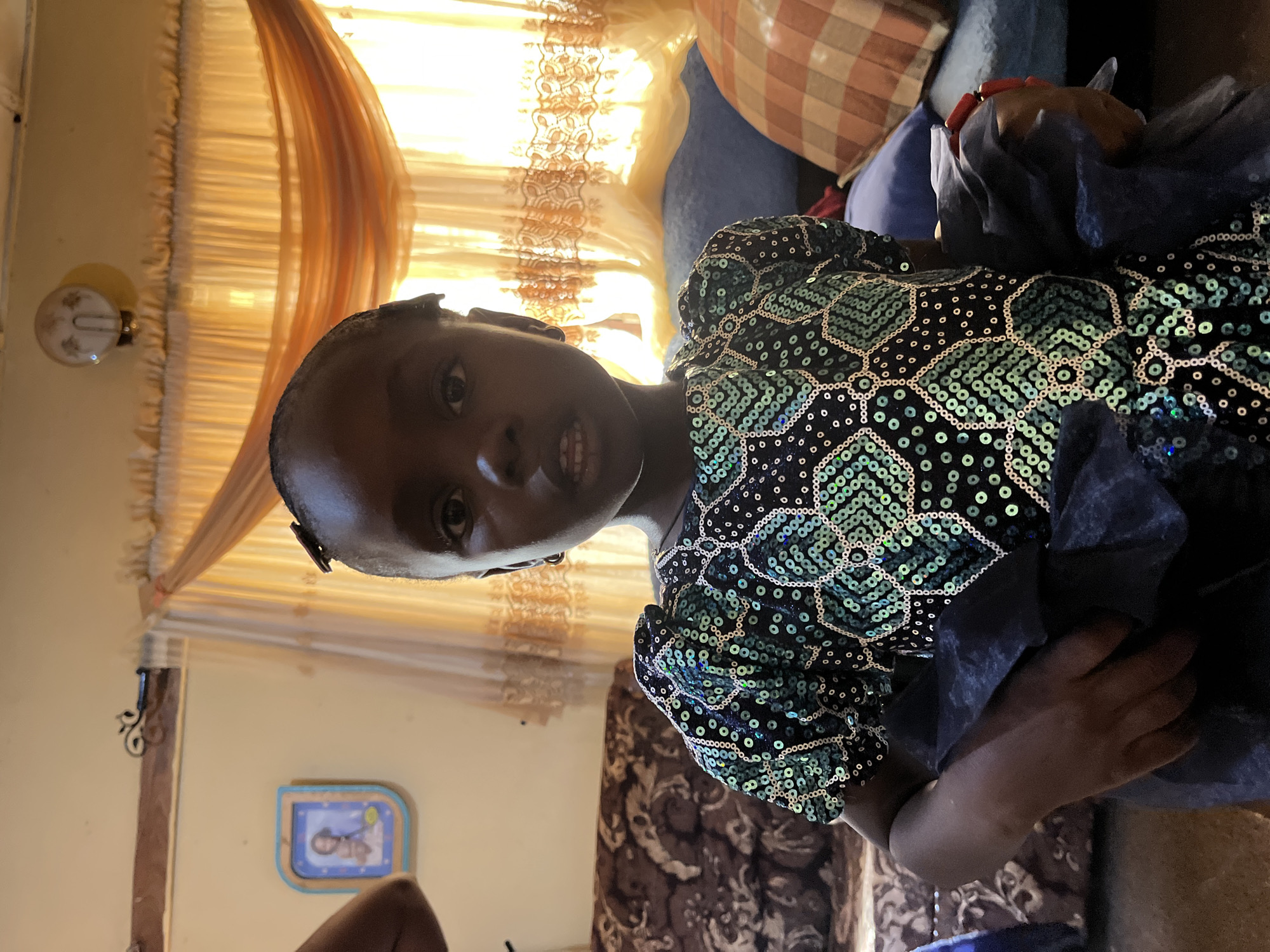 The image shows a young Black girl in a room in Rafin Bauna, Nigeria. She is wearing a black dress with geometric patterns of green sequins. The room has yellow walls and partially drawn sheer curtains. A small framed picture hangs on the wall. There is a sofa with patterned cushions and a blue pillow in the background. The girl is holding a dark blue piece of fabric.