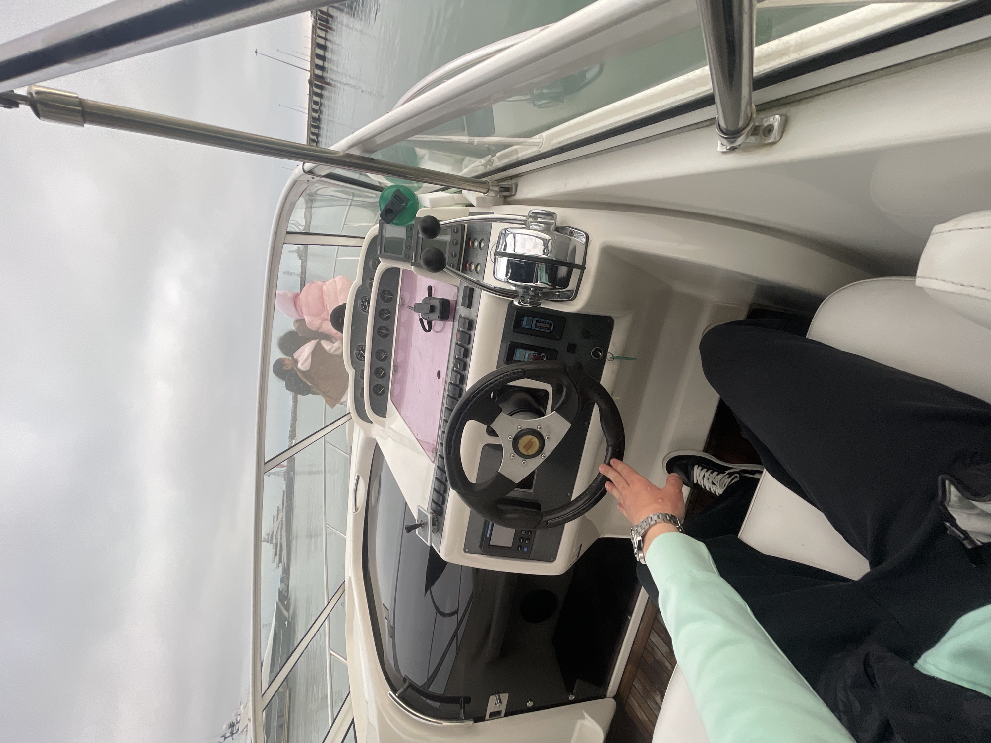 The image shows the interior of a boat's helm station, featuring a steering wheel, various gauges, and a throttle control. A person is seated, with one hand on the steering wheel and wearing a watch. Outside the boat's windshield, a cloudy sky and water are visible. There is a harbor or marina in the distance with masts of sailboats. Based on the context, this image was taken in Sochi, Russia.