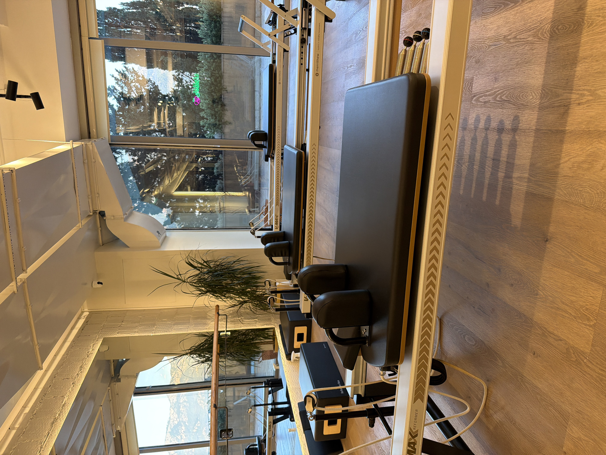 The image shows the interior of a pilates studio in Boreti, Montenegro. The studio is well-lit with natural light coming from large windows that overlook a green landscape with mountains in the distance. Several pilates reformers are arranged in the room, facing the windows. The reformers are black with wooden accents and have metal frames. There are also some black boxes with yellow accents, likely used for pilates exercises. Large plants are placed in pots near the windows, adding a touch of nature to the space. The floor is made of light-colored wooden planks, and the ceiling is white with exposed white beams and lighting fixtures. Mirrors are visible on the left side of the image, reflecting the studio space and the exterior view. There are no people visible in the image. The overall impression is one of a clean, modern, and tranquil fitness environment.