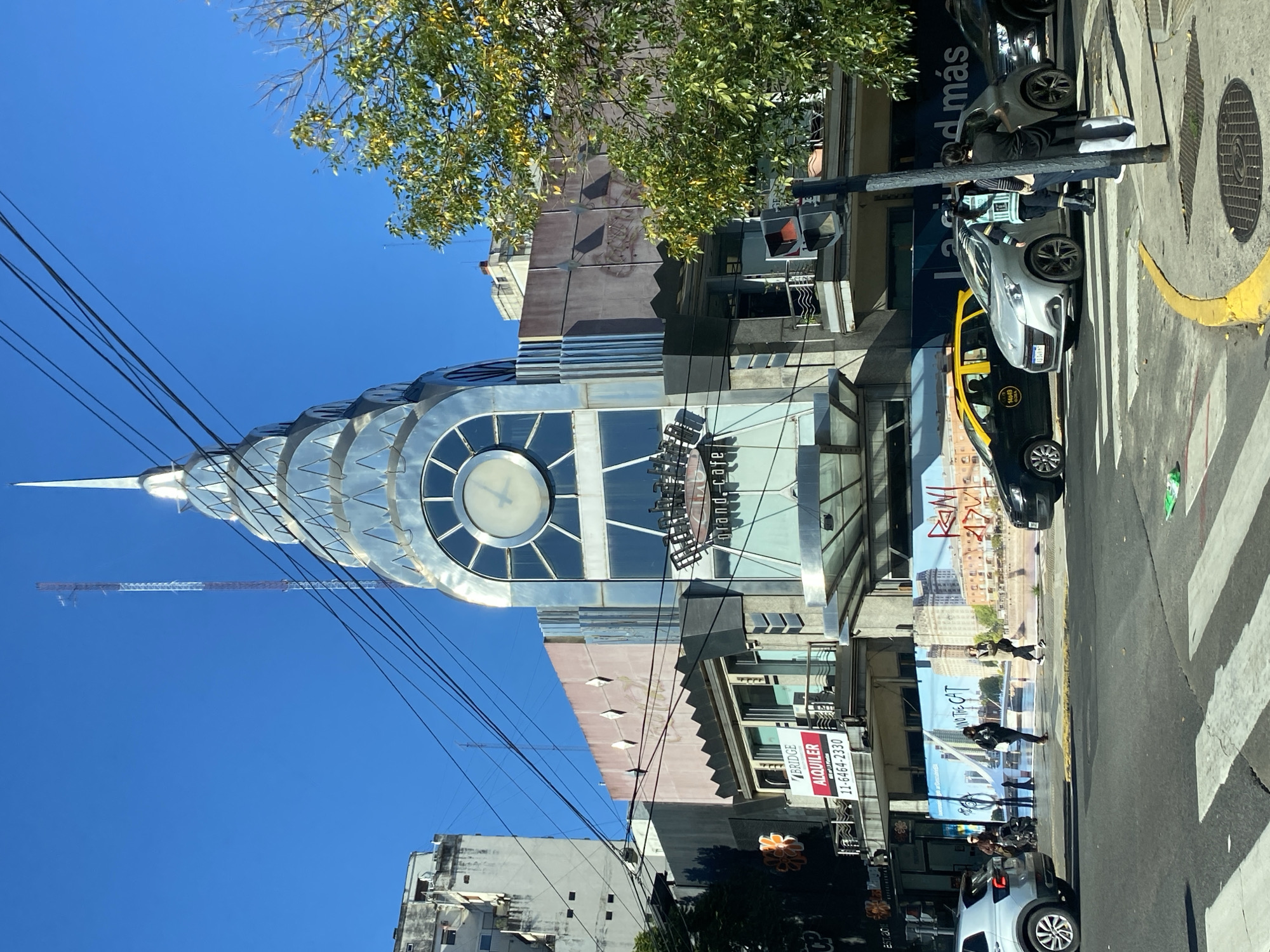 The image depicts a street scene in Buenos Aires, Argentina, featuring a building with a design resembling the Chrysler Building in New York. A prominent clock is integrated into the building's facade. Wires are visible across the sky, intersecting the building's spire. The street scene includes cars, a taxi, pedestrians crossing a crosswalk, and a street sign. The weather appears clear, with a blue sky. A construction crane is visible in the background. A sign advertises "Alquiler" with a phone number. A restaurant named "Gran Cafe" is also visible. A large advertisement with the words "ONLY THE CAT" is on the side of a building. The building's facade has a metallic finish.