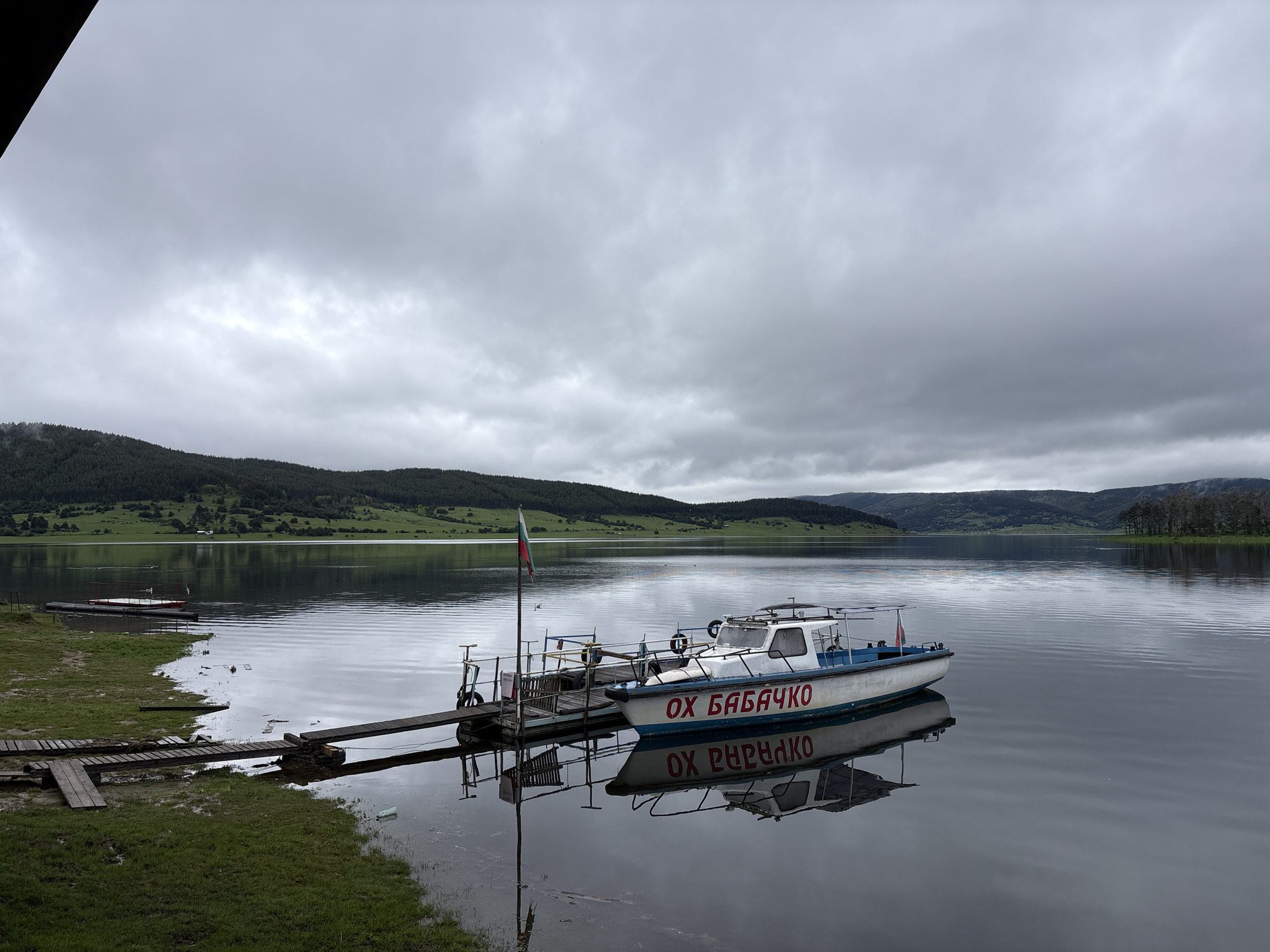 This image depicts a tranquil scene at Lake Batak in the city of Цигов чарк, Bulgaria. A small, white boat with blue trim and the inscription "ОХ БАБАЧКО" in red Cyrillic letters is docked at a wooden pier. A Bulgarian flag flutters from a pole on the pier. The still water reflects the boat and the overcast sky. Green hills line the horizon, and the overall environment suggests a calm, possibly cool, day. The pier extends from a grassy shoreline. The image captures a serene lakeside moment in this Bulgarian city.