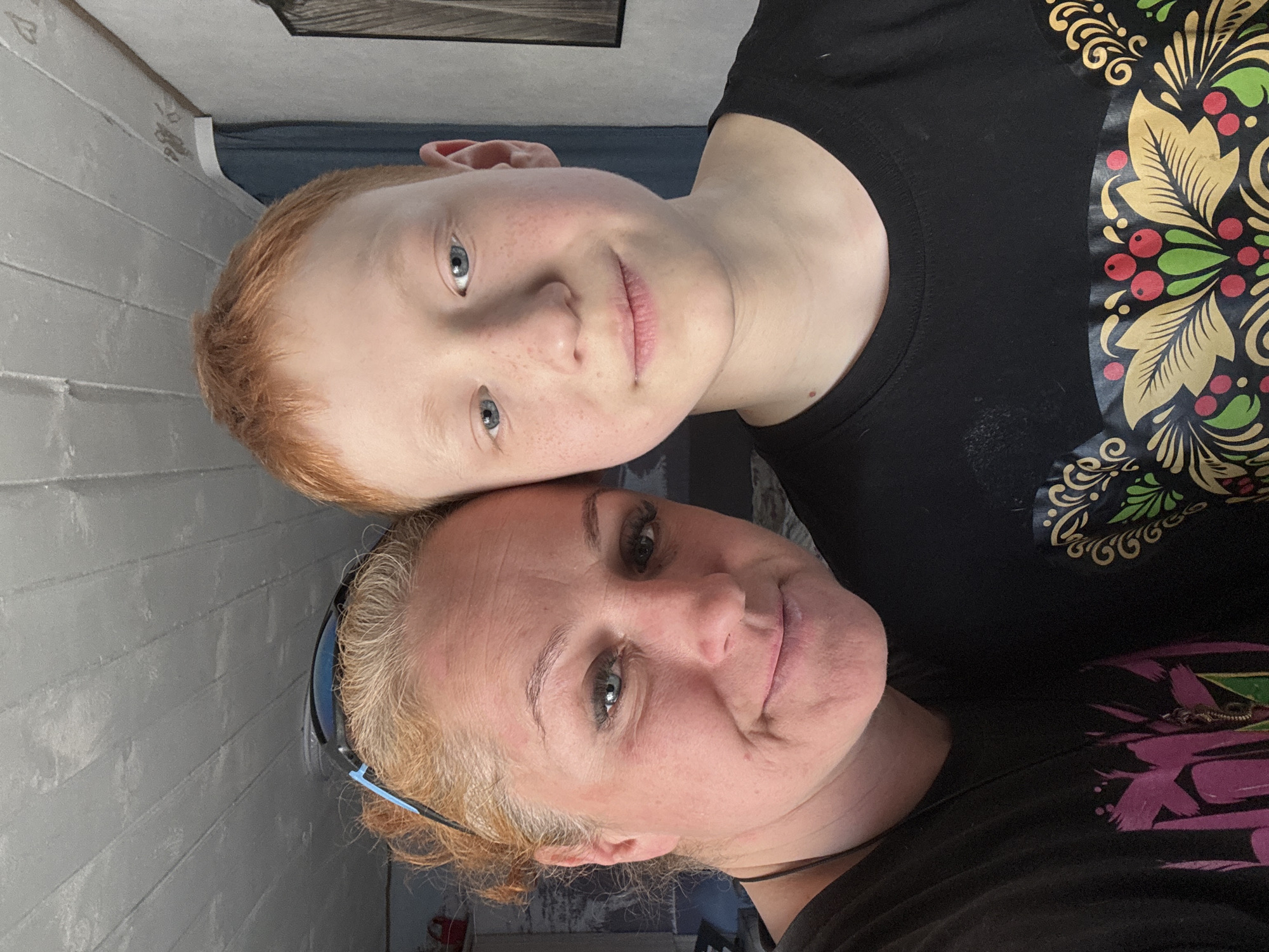 A casual, close-up portrait shows an adult woman and a boy, likely mother and son, affectionately posing with their heads together. The image is oriented vertically, with both subjects looking directly at the camera with gentle smiles, conveying a warm and happy mood.
The adult woman, positioned in the lower half of the frame, has light-colored hair, which is pulled back, and light blue or grey eyes. She has fair skin and is wearing blue-rimmed sunglasses pushed up onto her head. Her dark t-shirt features a colorful pattern with purple and green elements on the right side of the frame.
The boy, in the upper half of the frame, has distinctive reddish-blonde hair, fair skin with some freckles, and clear light blue eyes. He appears to be resting his head against the woman's. He is wearing a dark t-shirt adorned with a vibrant, folk-art inspired pattern featuring golden leaves, red berries, and green accents, reminiscent of traditional Russian decorative styles such as Khokhloma.
The background is softly blurred, suggesting an indoor setting. On the left side of the image (when oriented upright), a light-colored, possibly paneled or painted wooden wall is visible. No specific time of day or weather is discernible, and there is no visible text. The traditional design on the boy's shirt offers a subtle cultural cue consistent with the location context of Chaikovsky, Russia.