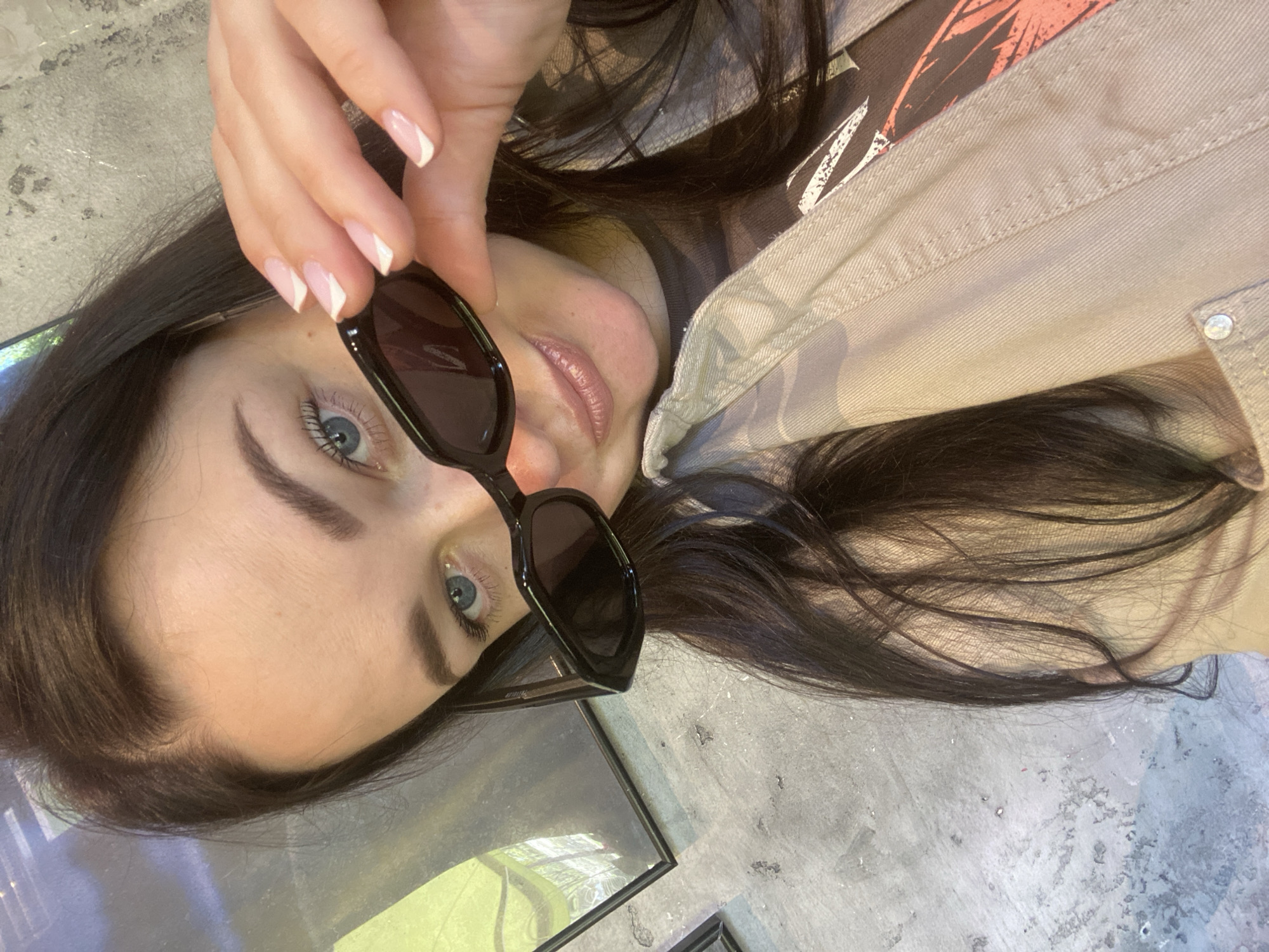 The image depicts a woman lying down, holding sunglasses in her hand. She has dark hair, blue eyes, and is wearing a tan jacket. The background is a gray, possibly concrete, surface. The location in Sochi, Russia cannot be determined from the information.