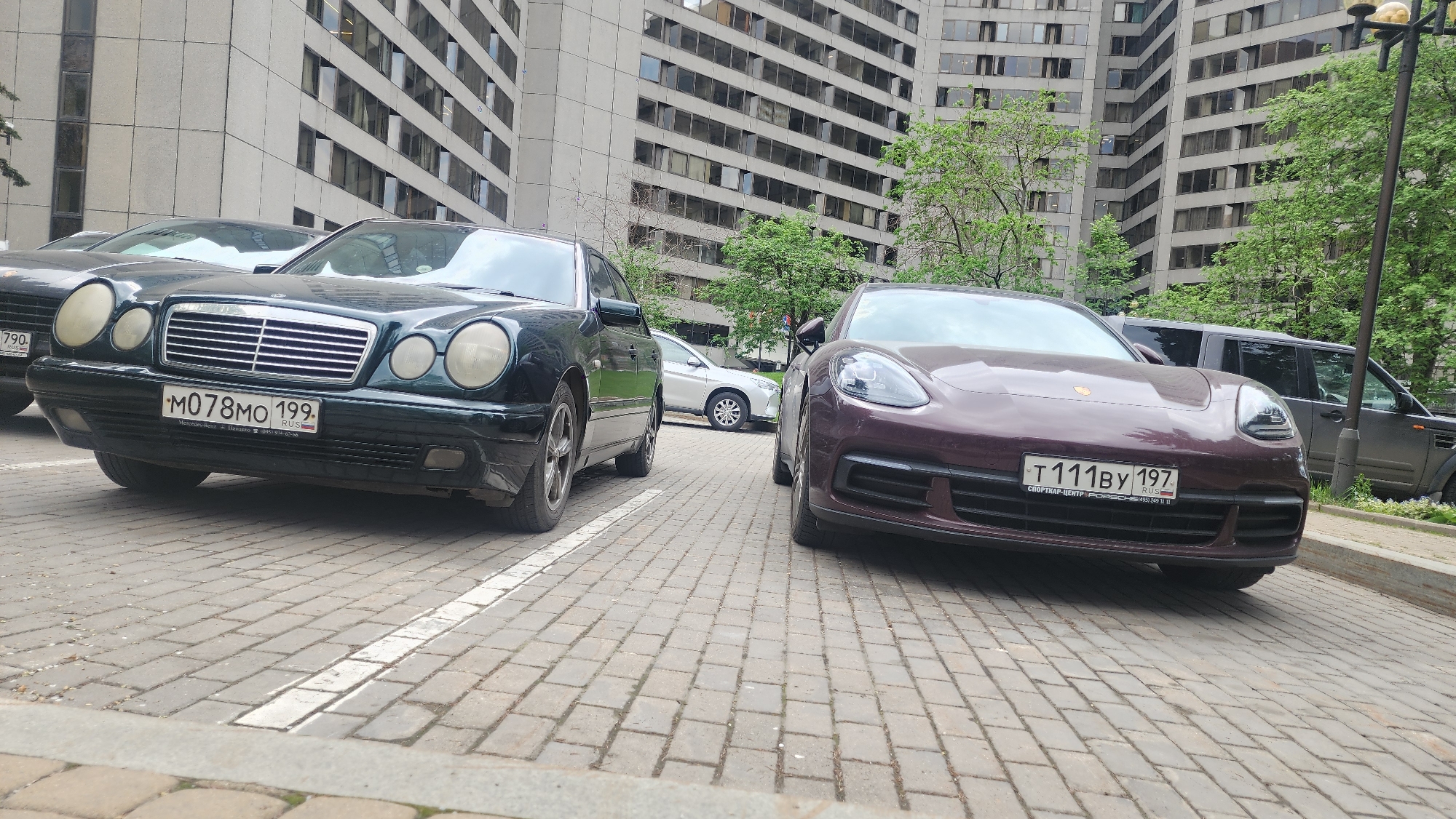 The image shows two luxury cars parked on a brick-paved area, likely a parking lot, in front of a large building. The car on the left is a dark green Mercedes-Benz E-Class (W210) with a license plate number "M078MO 199" from Moscow, Russia. The car on the right is a deep burgundy Porsche Panamera with a license plate number "T111BY 197", also from Russia. Other vehicles are visible in the background, including a dark SUV, a silver sedan, and a black car partially visible to the left of the Mercedes-Benz. The background building is a multi-story structure with numerous windows, typical of commercial or residential buildings in Moscow. Trees with green foliage are visible interspersed within the architectural landscape. A street lamp is visible in the background to the right. The paved area is marked with white lines, indicating parking spaces.