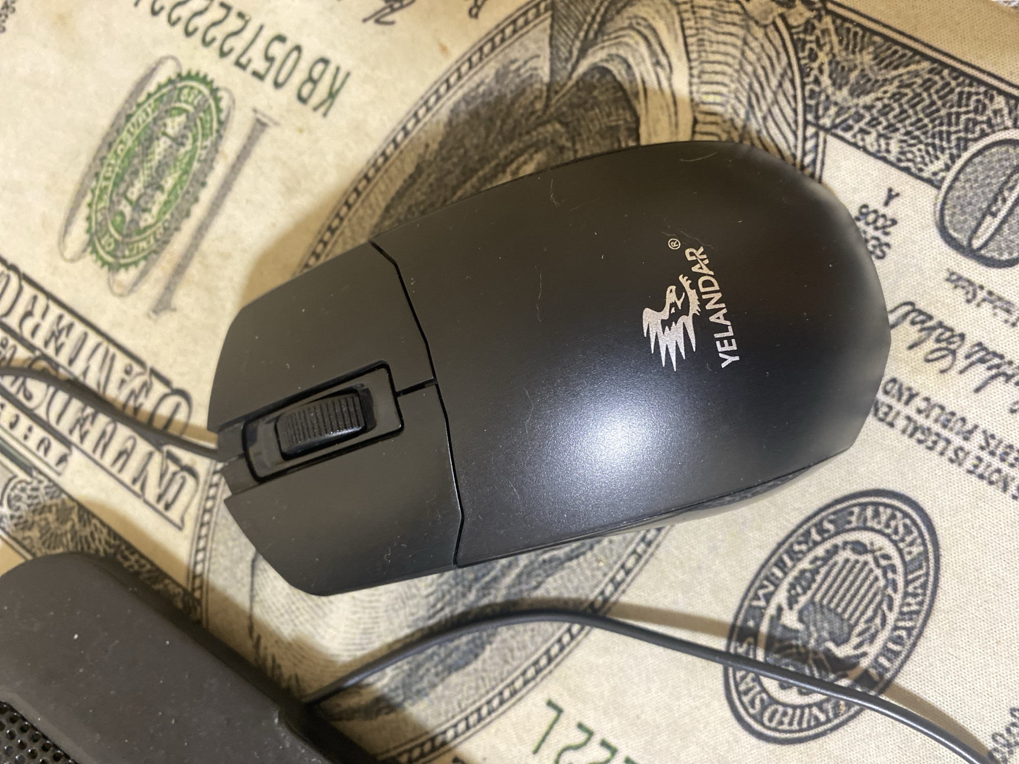 A black computer mouse with the "YELANDAR" logo is placed on a surface that appears to be a US dollar bill. The mouse is positioned in the center of the frame, angled slightly to the right. The scroll wheel is visible, along with the two main buttons. Part of another object, possibly a keyboard or a trackpad, is visible in the upper left corner of the image. The background is a textured surface with green and white markings that resemble currency. The scene is well-lit, with the focus primarily on the mouse. There are no people or animals visible in the image. The location in the city of Сонячне, Ukraine is not ascertainable from the provided image.