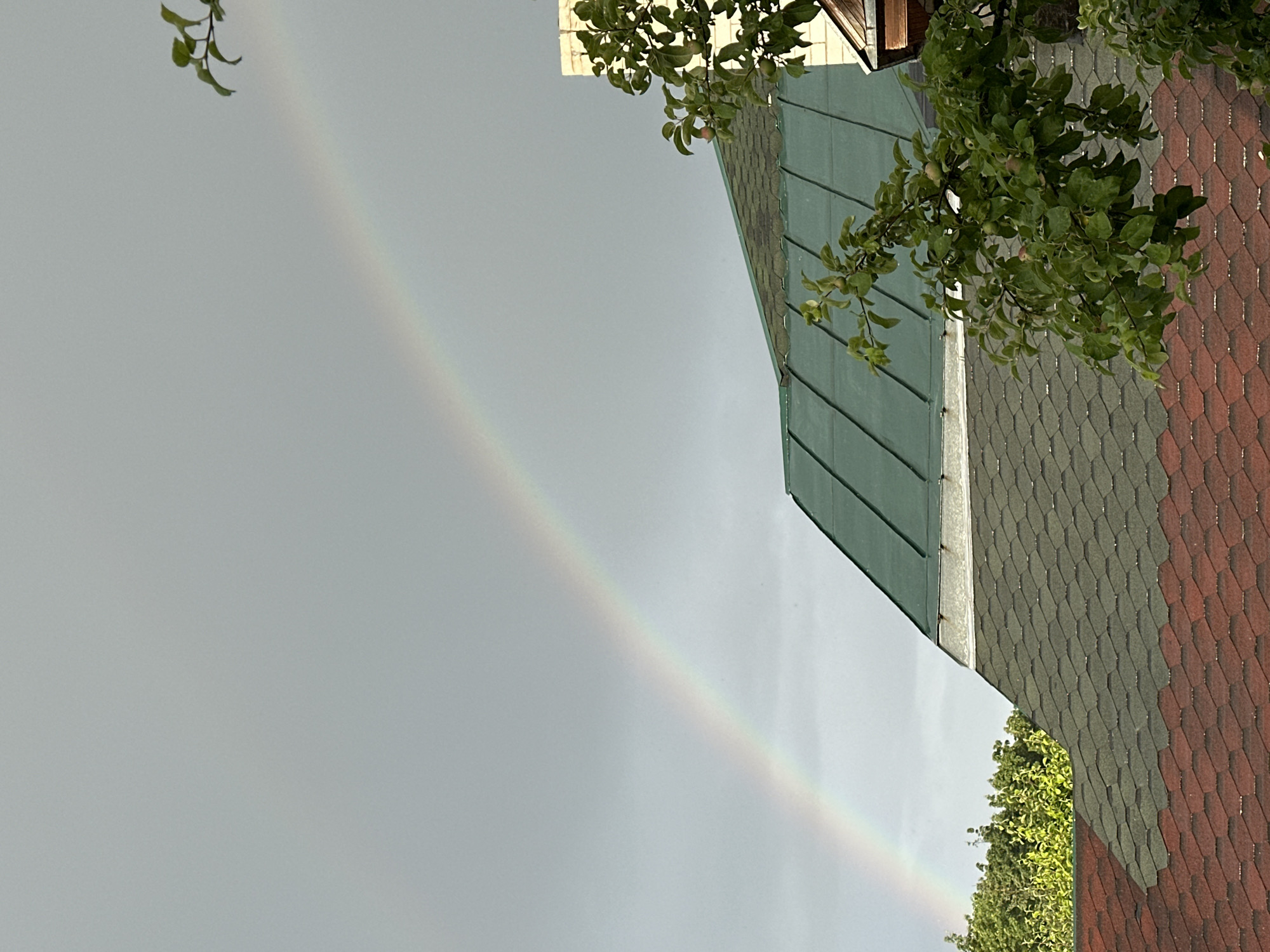 The image captures a view of a residential area in Nizhny Novgorod, Russia, featuring a rainbow stretching across a cloudy, overcast sky. The foreground is dominated by the angled roofs of houses, one with dark green shingles and another with reddish-brown shingles, creating a textured visual contrast. Lush green foliage from trees partially obscures the roofs and the sky, suggesting it is either spring or summer.
The rainbow, a subtle arc of pastel colors, begins in the upper left quadrant of the image and extends diagonally towards the lower right. Its faint appearance suggests it may be a secondary rainbow or the sun is not fully out. The overall lighting is diffused, indicating a cloudy or post-rainy weather condition. There are no people visible in the frame, and the focus is on the natural phenomenon and the architectural elements of the houses. The scene evokes a sense of calm and natural beauty, characteristic of a quiet day in a suburban or residential setting. No text is visible in the image.