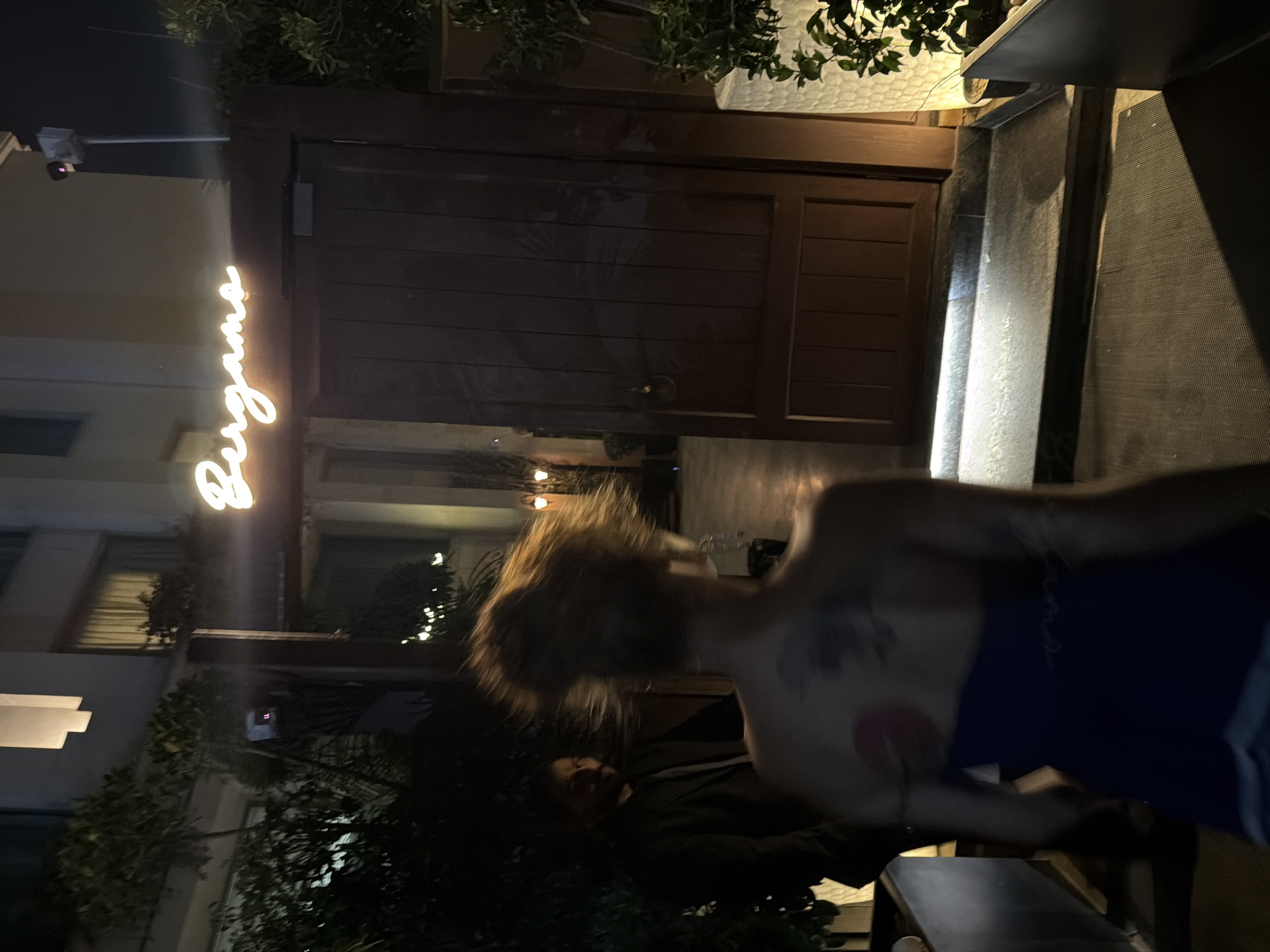 The image depicts an outdoor scene at night, possibly in New Delhi, India, with the illuminated sign "Bergamo" above a dark wooden door. In the foreground, a person with reddish-brown hair is seen from behind, wearing a dark blue strapless dress. They have tattoos on their upper back, including a red circular design on the left shoulder blade and a more complex tattoo on the right. To the left of this person, a man in a dark suit and tie is visible, looking towards the camera. He is partially obscured by foliage. The environment features some greenery and lighting fixtures.