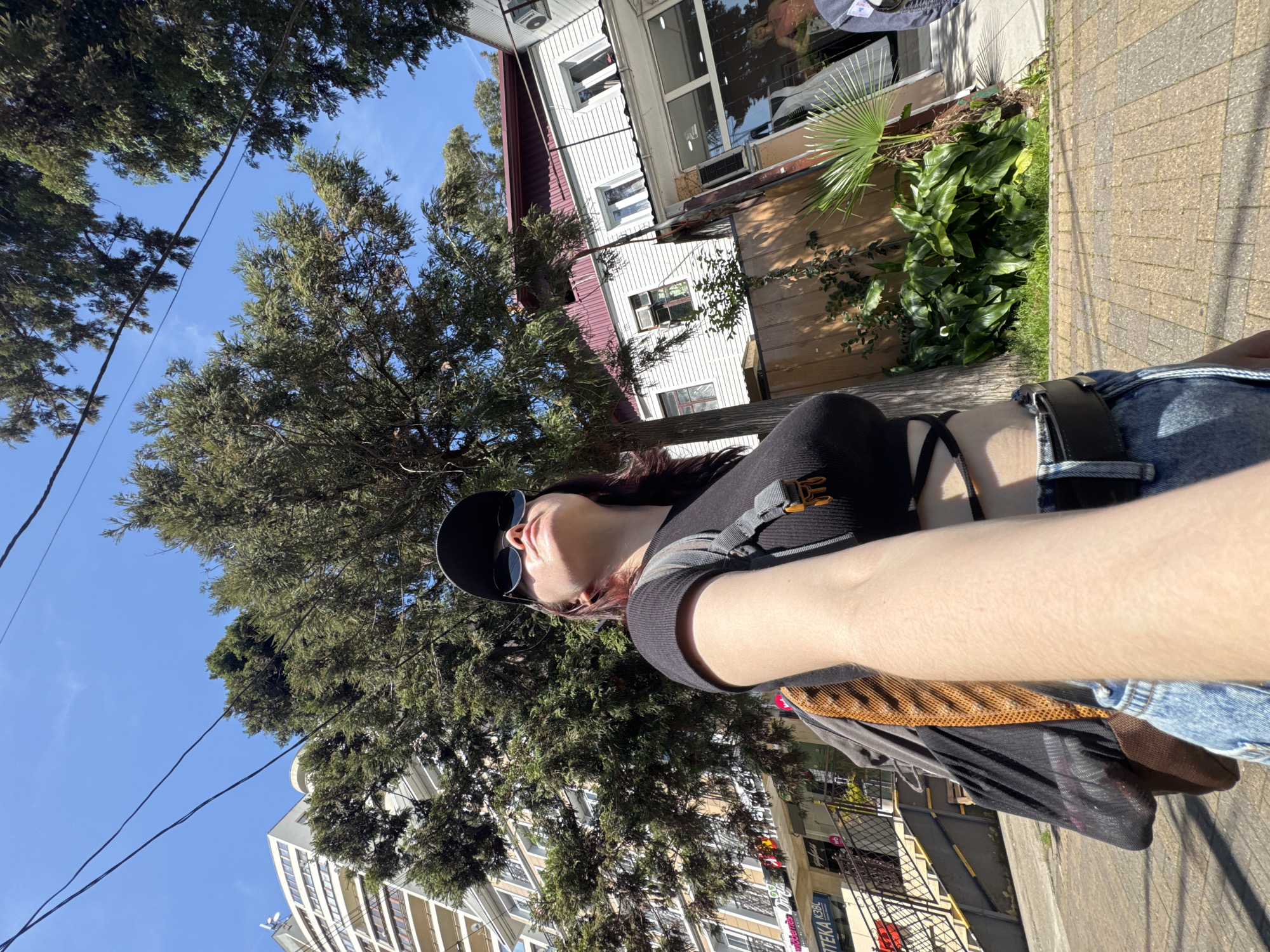 A person wearing a black t-shirt, sunglasses, and a cap is taking a selfie outdoors. The background includes buildings, trees, and a clear blue sky. The location is Sochi, Russia.