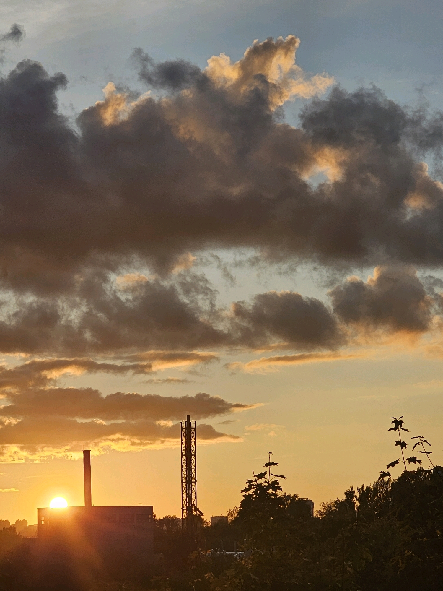 The image depicts a sunset over the city of Khimki, Russia, viewed from a slightly elevated position. The sky is filled with dramatic clouds, some dark and heavy, others illuminated by the setting sun with hues of orange and yellow. In the foreground, silhouetted trees and foliage frame the lower portion of the image.
Dominating the middle ground are industrial structures. To the left, a large, rectangular building with a chimney stands prominently. To its right, a tall, lattice-like tower, possibly an industrial chimney or communication mast, rises into the sky. The sun, a bright orb, is positioned behind the building on the left, casting a warm, golden glow across the scene and creating lens flare. In the distance, faint outlines of other buildings and trees can be seen.
No people are visible in the image. The scene is static, capturing a moment of natural beauty intersecting with urban industrial landscape. The lighting suggests it is late afternoon or early evening.