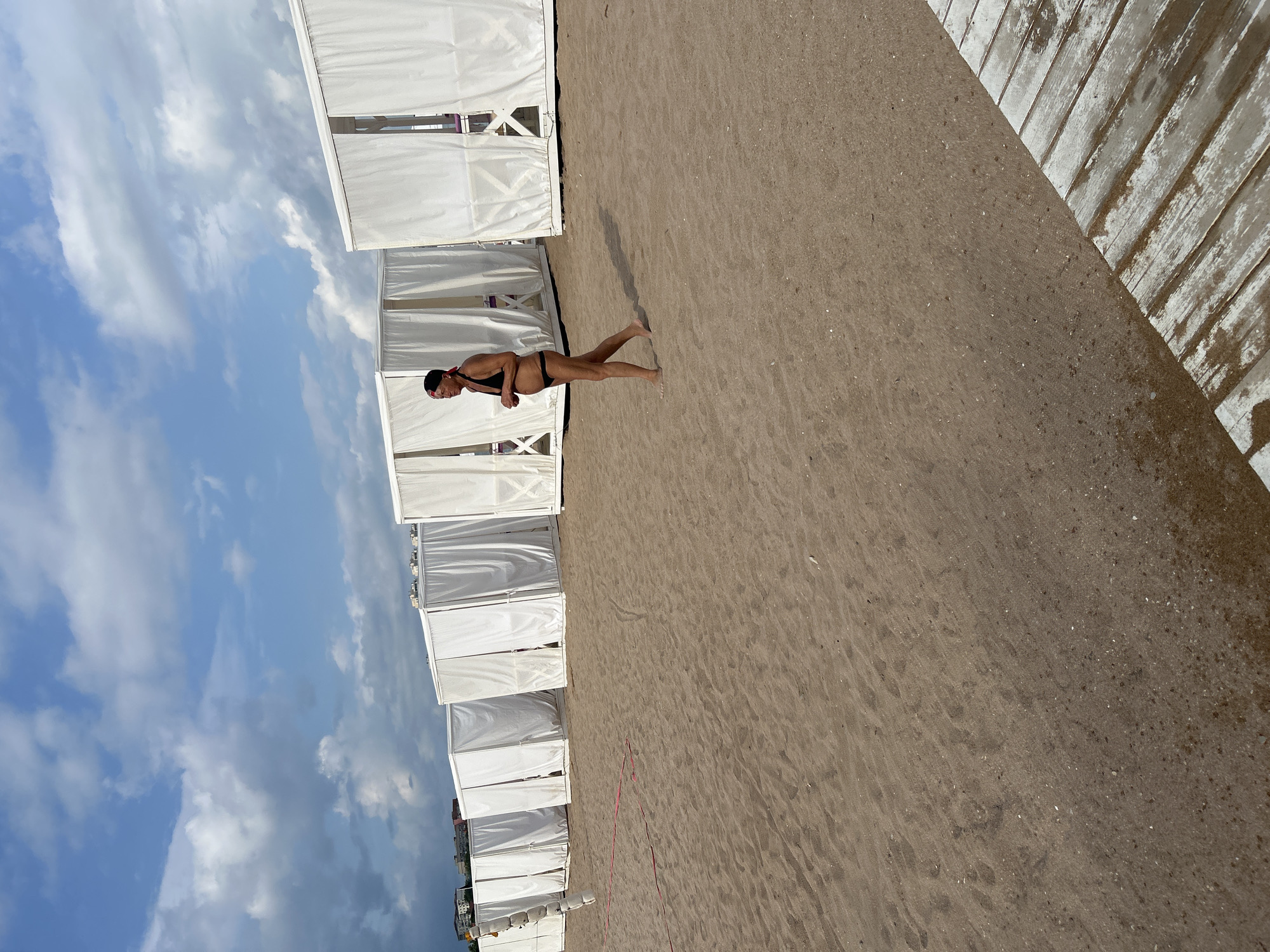 A woman wearing a black bikini walks on a sandy beach lined with white cabanas. The cabanas, with their curtains drawn, stretch into the background. The sky above is blue with scattered clouds. In the foreground, a wooden walkway is visible on the right side of the image. This scene is located on the beach in Gelendzhik, Russia.