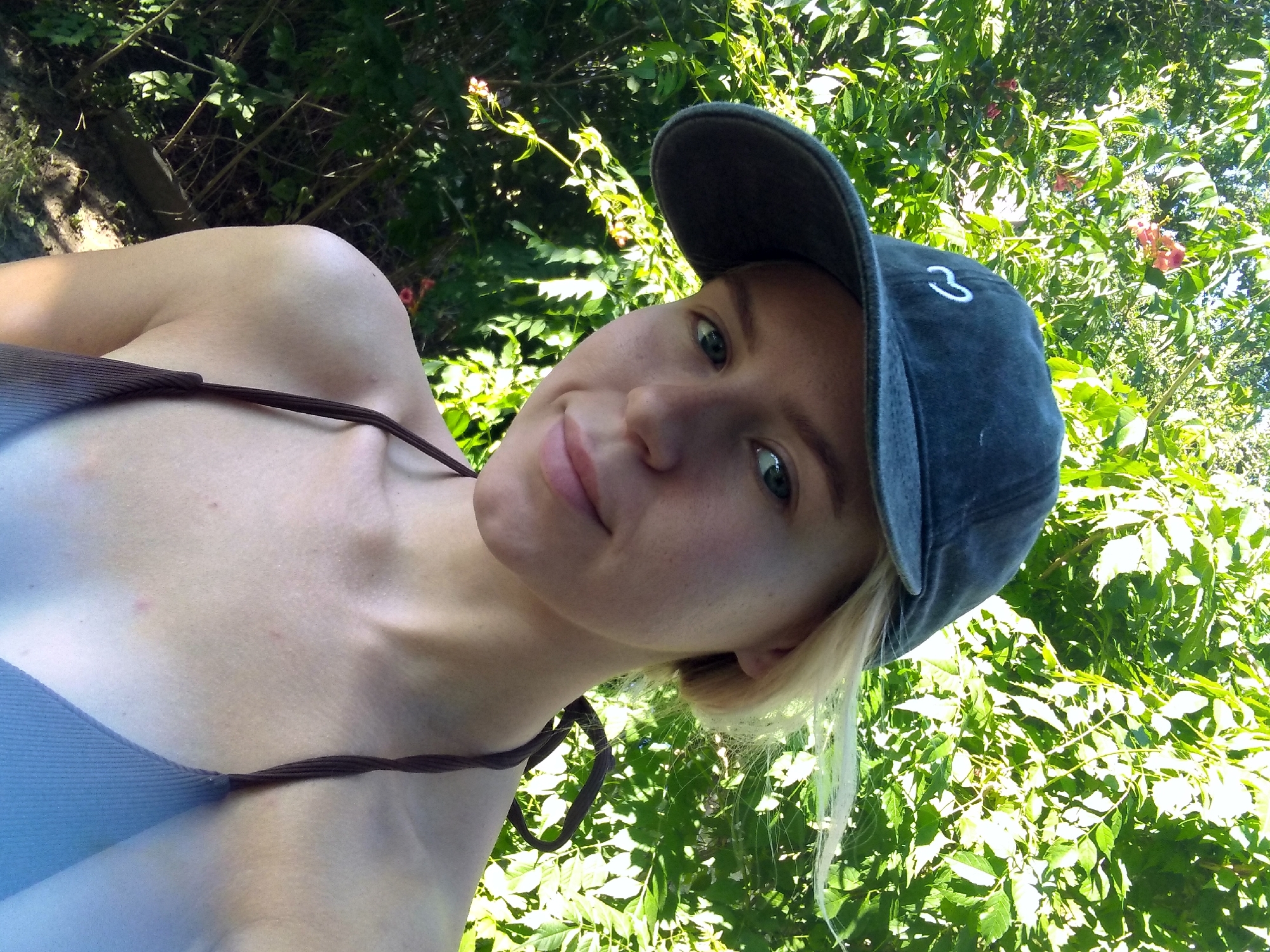 A young woman with blonde hair, wearing a dark blue baseball cap and a brown bikini top, is photographed outdoors. She is looking towards the camera with a slight smile. The background is filled with lush green foliage, suggesting a natural setting. The lighting indicates it is daytime, with sunlight filtering through the leaves. The location is identified as Radisne, Ukraine.