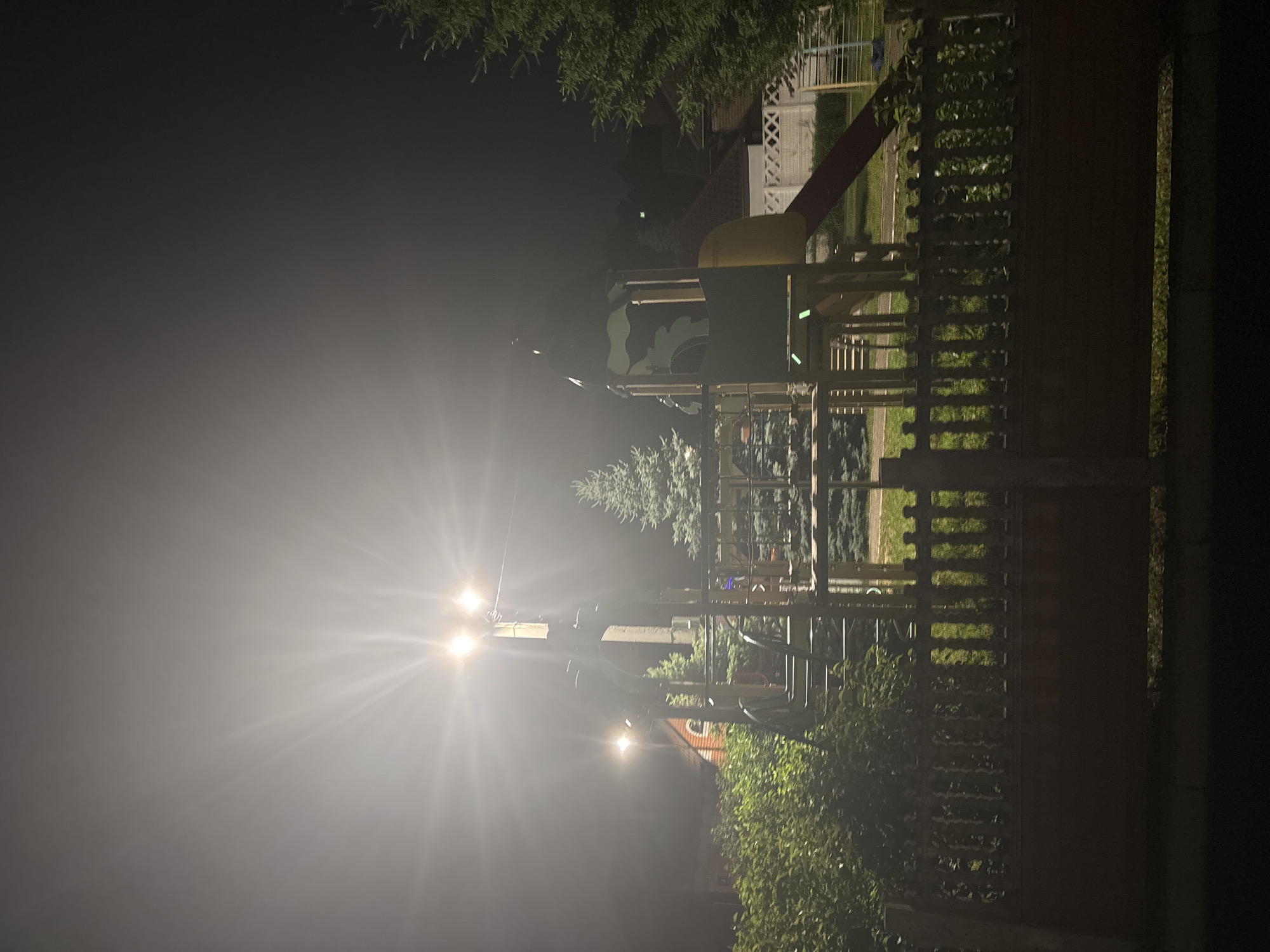 The image depicts an outdoor playground at night, illuminated by bright streetlights. The playground features a green and yellow climbing structure with a red slide. A wooden fence is visible in the foreground, with lush green bushes behind it. A tall evergreen tree stands to the right of the playground. In the background, a red-brick house with arched windows is visible, as is a white lattice fence. The scene is set in the city of Orekhovka, Russia.