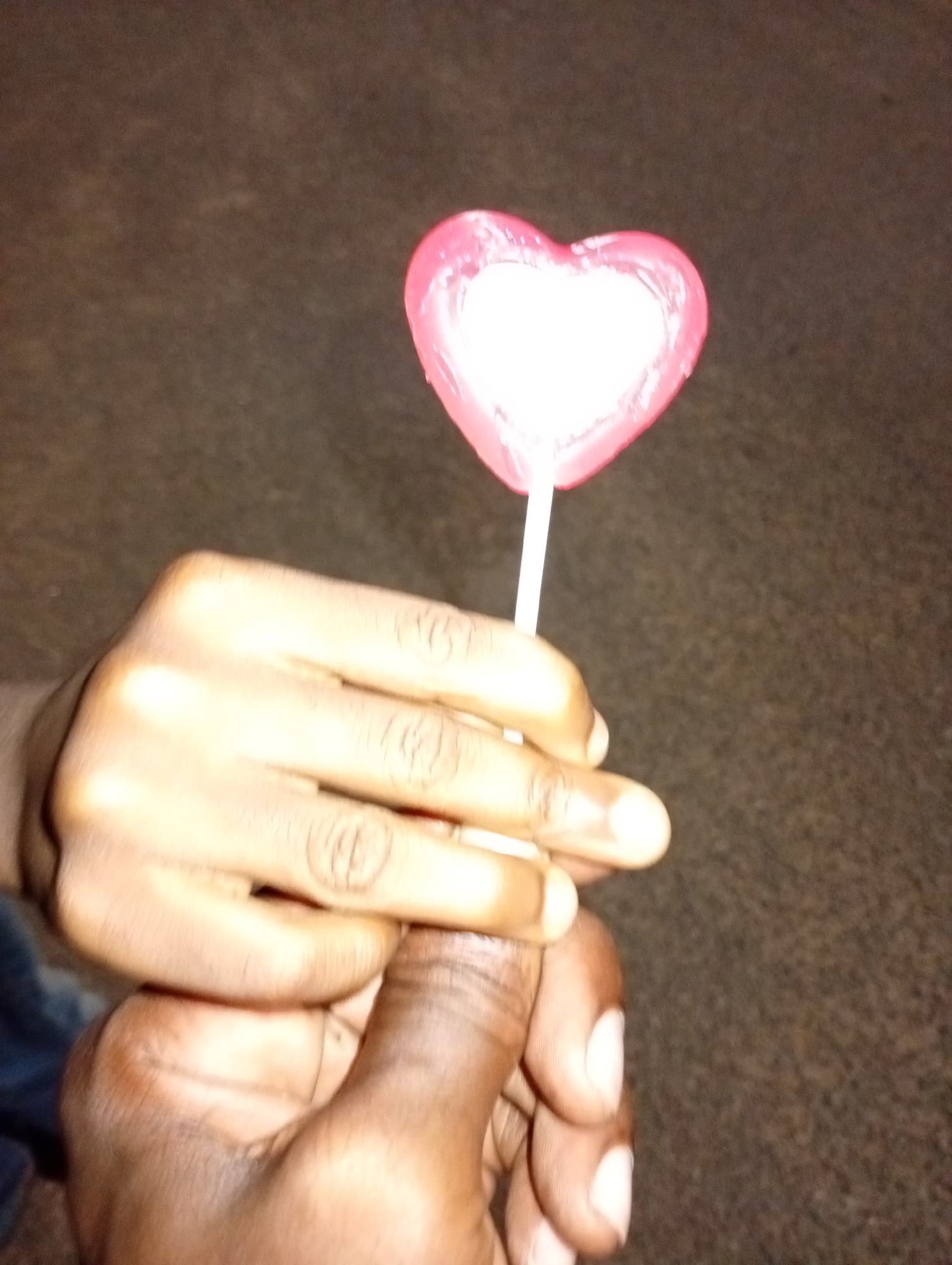 Two hands are holding a heart-shaped lollipop against a blurry, dark brown background. One hand is darker-skinned and appears to be holding the lollipop stick, while a lighter-skinned hand is wrapped around it. The lollipop is pink with a white center. The location is not discernible from the image.