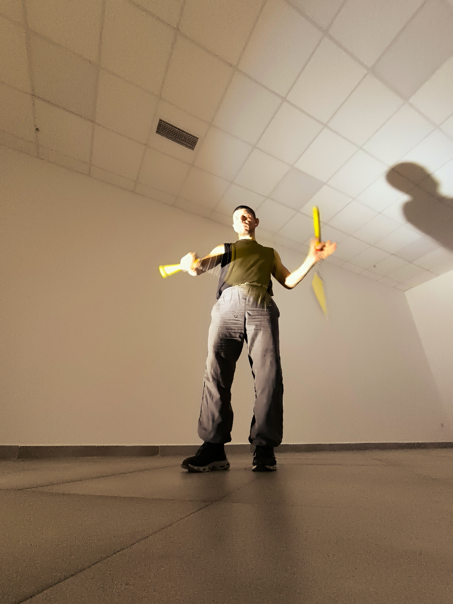 A person is standing in a room with a drop ceiling and white walls, holding two yellow objects, possibly nunchucks or juggling clubs. The person is wearing a dark olive-green sleeveless top, dark gray baggy pants, and black athletic shoes. The lighting in the room is dim, casting a shadow of the person on the ceiling. The floor appears to be tiled. The image is taken from a low angle, looking up towards the person and the ceiling. The overall atmosphere is somewhat stark and minimalist.