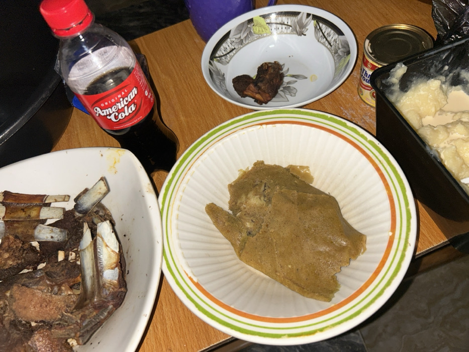 In the city of Debasco, Nigeria, a wooden table displays a prepared meal. A large white oval platter at the upper left holds multiple pieces of dark brown, roasted rib meat. Below this, a white bowl with green and orange stripes contains a substantial portion of light brown, mashed food. To the right, a small white bowl with a floral pattern holds a single piece of dark brown roasted meat. A partially compressed clear plastic bottle of "Original American Cola" with a red cap is positioned near the roasted ribs. In the lower right, a black container partially reveals a creamy, off-white, lumpy substance, with a metal can visible next to it. No individuals are present in the scene.