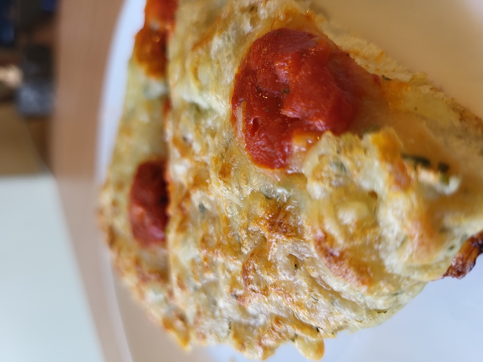 A close-up, shallow depth of field shot shows a slice of pizza on a white plate. The pizza has a golden-brown crust, melted white cheese, and dollops of red sauce. Some green herbs are visible mixed into the cheese. The background is blurred, with a wooden surface and a hint of a dark object. The location is unknown, as no identifying landmarks are visible.