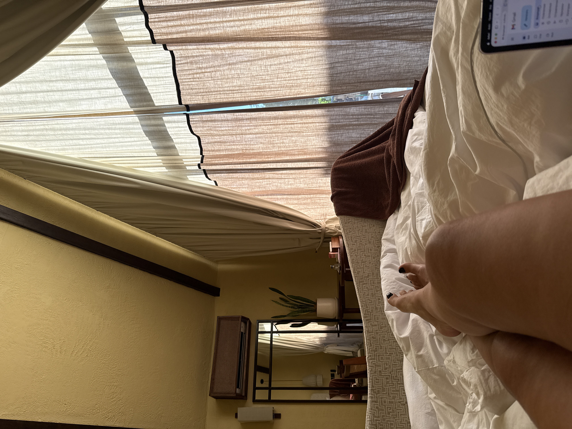 The image captures a first-person perspective from a bed in what appears to be a bedroom, likely in a hotel or guesthouse in Kuta Selatan, Indonesia. The overall scene conveys relaxation during daytime.
The primary subject is the room's interior. In the foreground, the lower right shows a person's legs and feet with black painted toenails, resting on white bedding, which consists of sheets and a duvet. A dark brown textured blanket is draped over the bed on the left side. The person's hand is visible, holding a smartphone that displays a blurred screen, where the Gmail logo can be faintly discerned, indicating email or messaging activity.
The room features a large window covered by layered blinds or shades in beige and light brown tones. Bright sunlight streams through the blinds, casting distinct shadows and illuminating the room. A narrow gap in the blinds reveals a sliver of the outdoor environment with green foliage, suggesting a sunny day.
The walls are textured and painted a light yellow or ochre color, complemented by dark brown wooden trim running along the top edges. On the wall to the left, there's a rectangular mirror with a black metal frame, reflecting parts of the room, including the blinds and a snake plant (Sansevieria) in a white pot. Below the mirror, a small white roll (possibly toilet paper) is mounted on the wall. To the left of the mirror, a brown woven rectangular unit, possibly a shelf or storage box, is affixed to the wall. The general decor with its natural tones and wooden elements aligns with a tropical or resort setting.