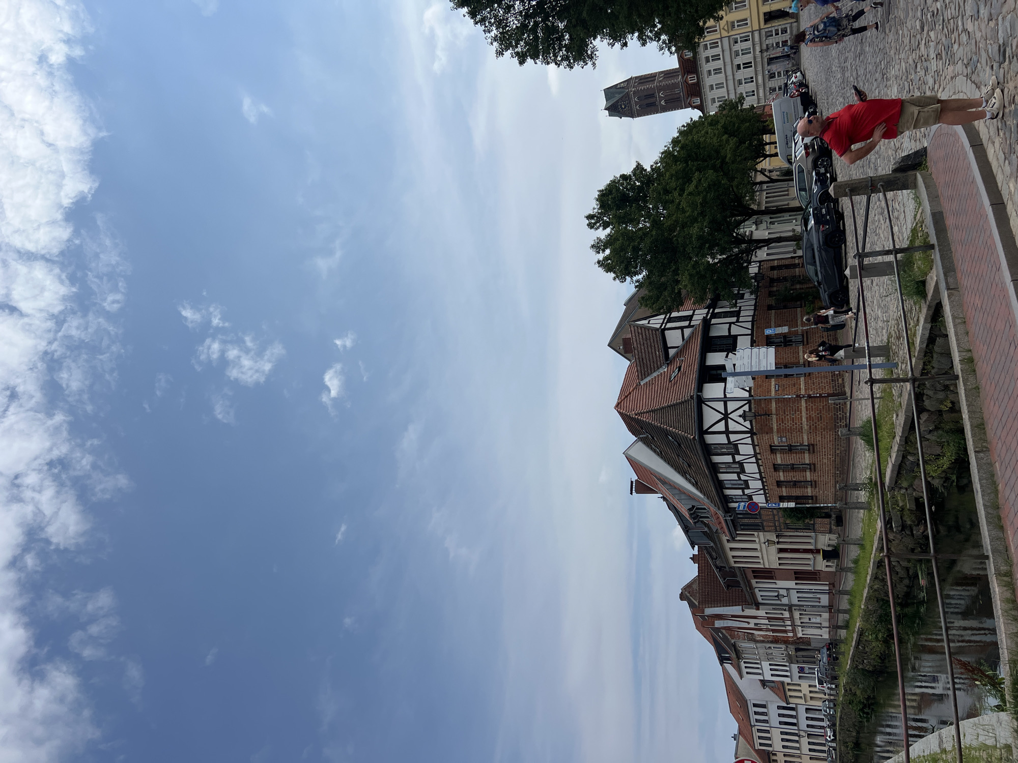 The image shows a street scene in Wismar, Germany. In the foreground, a man in a red polo shirt and khaki shorts stands with his hands on his hips, holding a phone. He is on a cobblestone street. Behind him, there is a canal with a stone embankment and a metal railing. On the other side of the canal are several buildings, including a half-timbered house with red tile roofs. Further down the street, a tall church tower is visible. Several cars are parked along the street. In the background, there are more buildings and trees. The sky is blue with scattered white clouds.