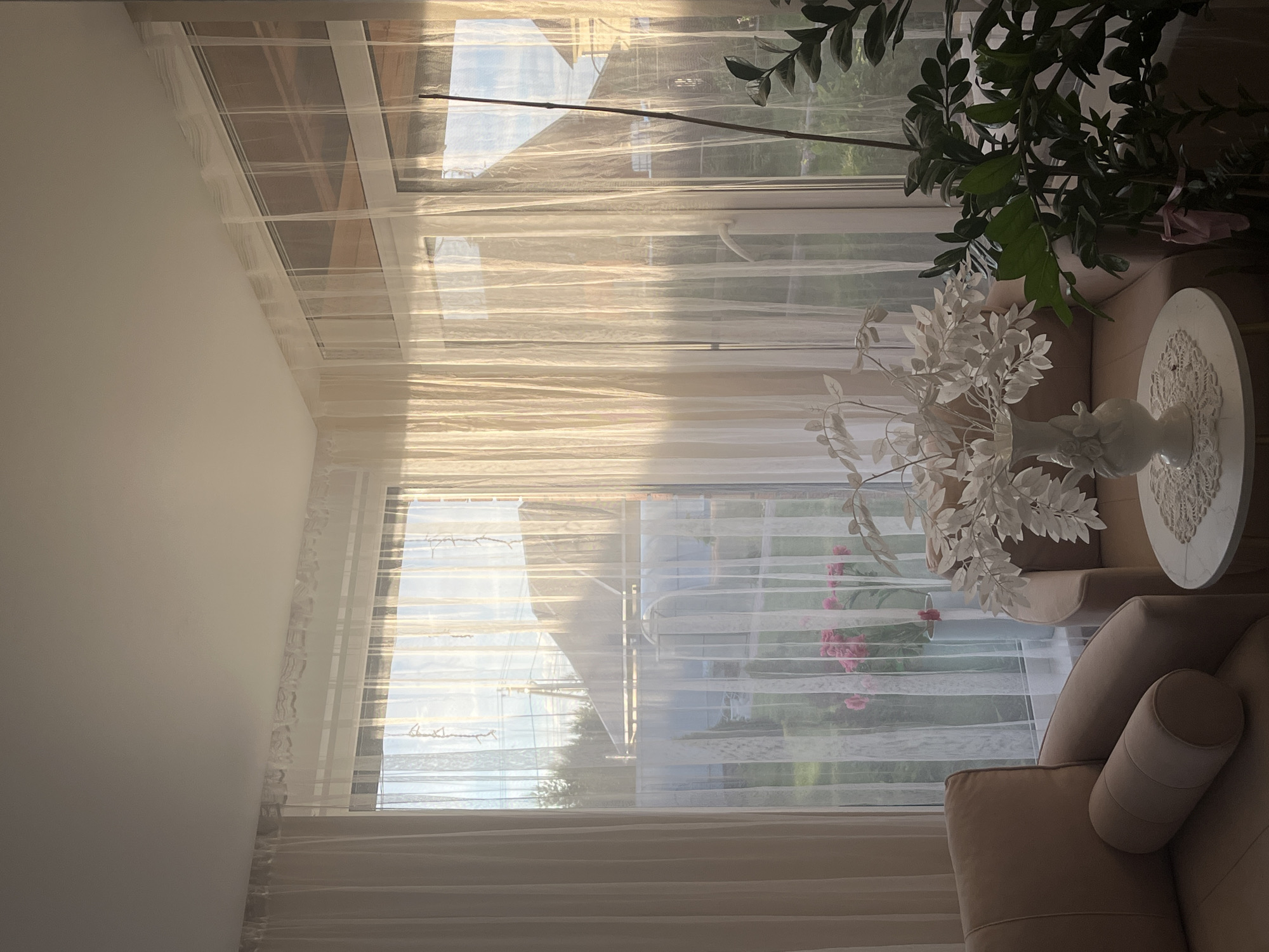 The image depicts an interior scene in what appears to be a room with a large window. Sheer white curtains filter the sunlight, creating a soft, diffused light in the room. Outside the window, there is a view of trees and residential buildings, possibly in a suburban or residential area.
Inside, a tan-colored sofa is visible, with a rolled cushion on one end. A round white table sits beside the sofa, adorned with a vase of white artificial flowers and a lace doily. A leafy green plant stands nearby, partially obscuring the view. The room is brightly lit, suggesting it's daytime. The location is not possible to determine with certainty, even knowing it's in Chaikovsky, Russia, but it appears to be a living space or a room intended for relaxation and comfort.