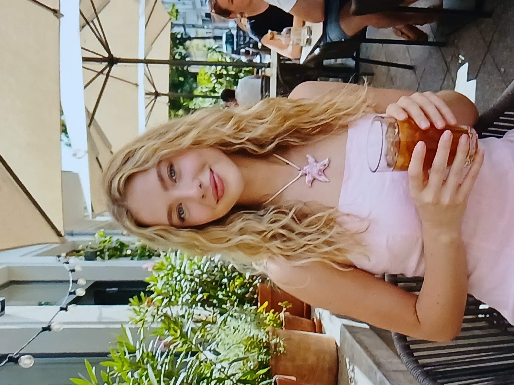 A young woman with long blonde, wavy hair and a pink starfish pendant is seated at an outdoor cafe, holding a glass of iced tea. She is wearing a light pink strapless dress. She is looking at the camera and smiling. In the background, other patrons are seated at tables under umbrellas, and there are potted plants along a wall. The setting appears to be a street-level cafe with a relaxed atmosphere. There is no specific indication of the location being the city of Dnipro, Ukraine.