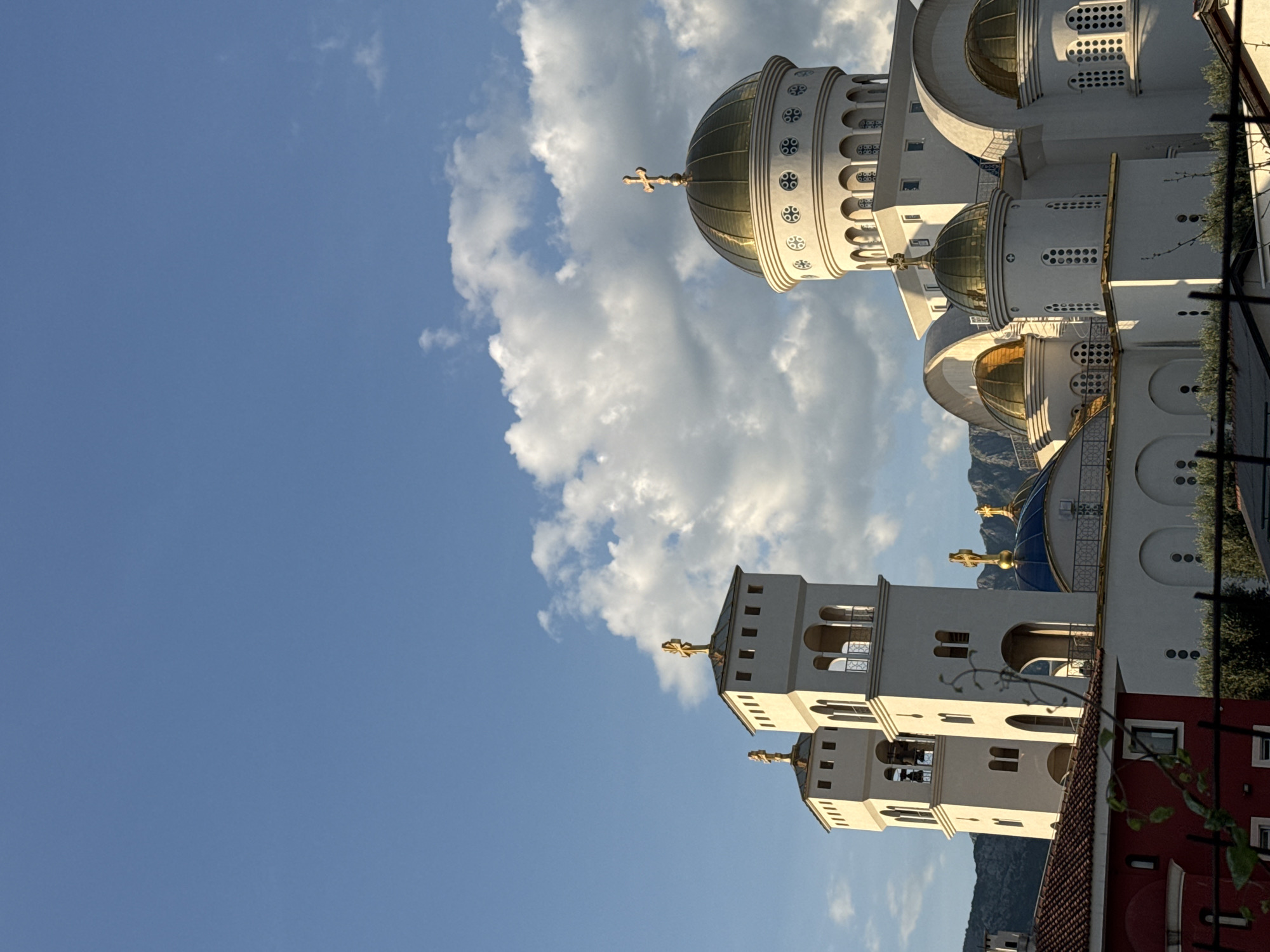 The image shows the exterior of the St. John Vladimir's Church in Bar, Montenegro. The church features white walls, golden domes, and two bell towers, with ornate designs and crosses atop its structures. The sky is blue with scattered clouds. Part of a red building is visible in the foreground. Mountains are visible in the background.